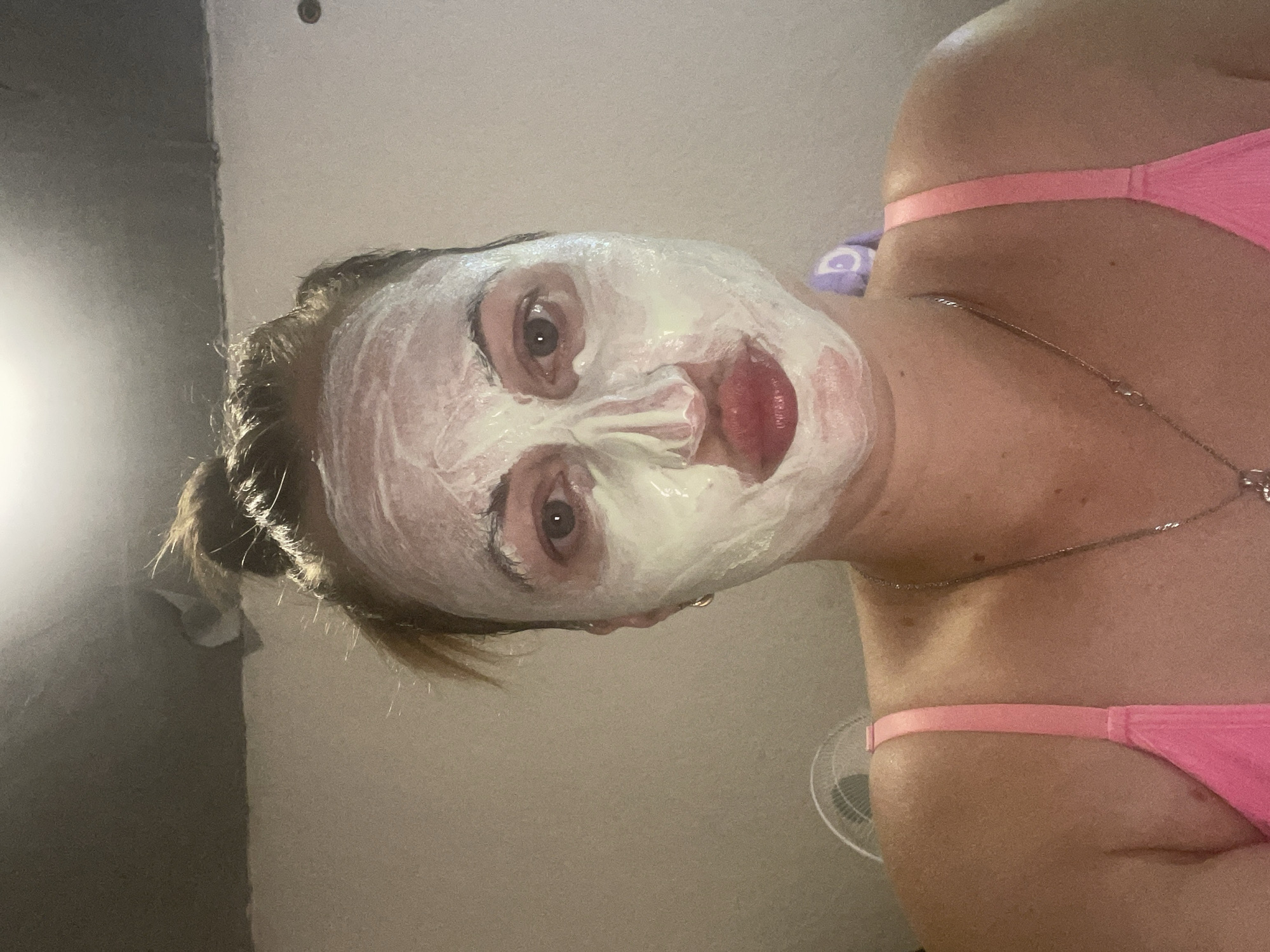 A woman with blonde hair pulled up in a messy bun is shown from the chest up. She is wearing a pink bra and a silver necklace. Her face is covered in a white and green face mask. She is looking directly at the camera with her lips slightly parted. The background appears to be a plain wall with some ceiling detail. There is a fan visible to the left of the woman. The image does not provide any information about the location being in Zarichne, Ukraine.