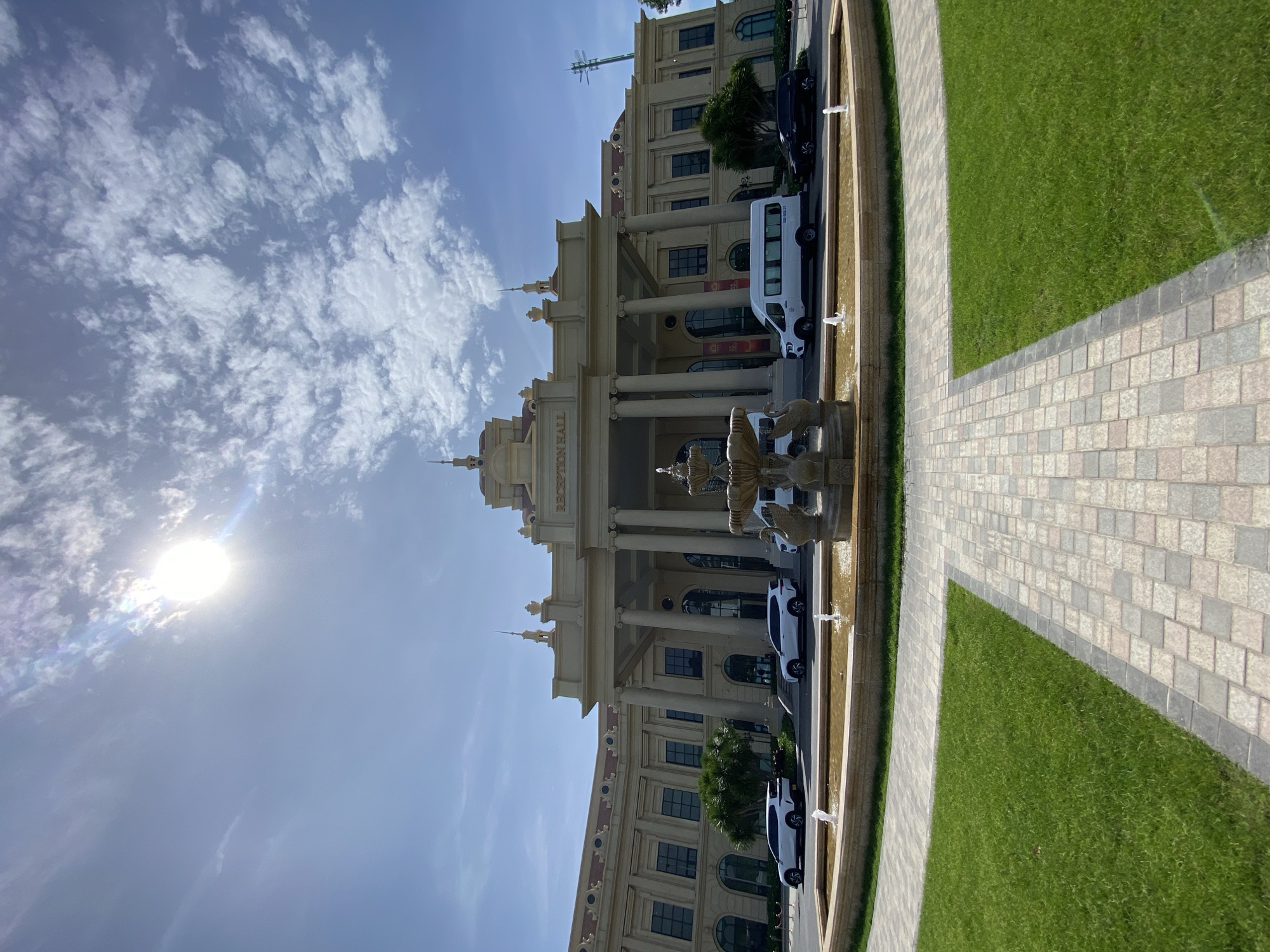 The image shows the entrance of a building labeled "RECEPTION HALL" in Nha Trang, Vietnam. The building has a grand, classical architectural style with large columns and a curved facade. In front of the building, there is a circular fountain with water jets. A paved walkway leads from the foreground towards the fountain and the building. Well-maintained green grass surrounds the walkway and fountain. Several vehicles, including white SUVs, a white van, and a black SUV, are parked in front of the building. The sky is blue with scattered white clouds, and the sun is bright, creating some lens flare in the upper left portion of the image.