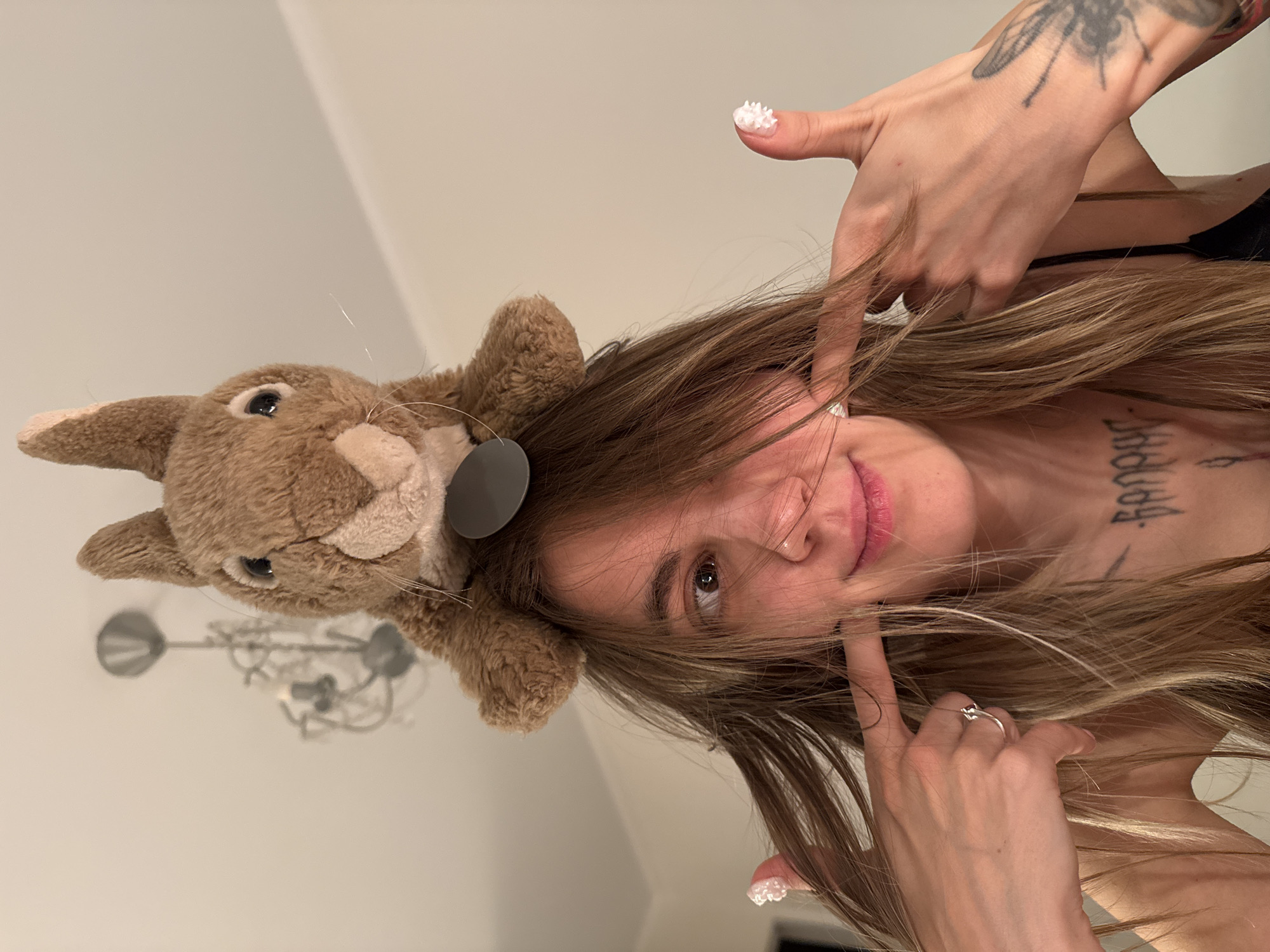 A young woman with blonde hair and a stuffed rabbit toy on her head is posing for a selfie. She is wearing a black tank top and has tattoos on her chest and arm. Her fingernails are painted white and appear to be decorated with small white embellishments. She is using her fingers to pull at her cheeks, creating a playful expression. The background appears to be an indoor setting with a chandelier visible on the ceiling. The lighting is warm and suggests an evening or indoor environment. The image does not provide any information about the location being Moscow, Russia.