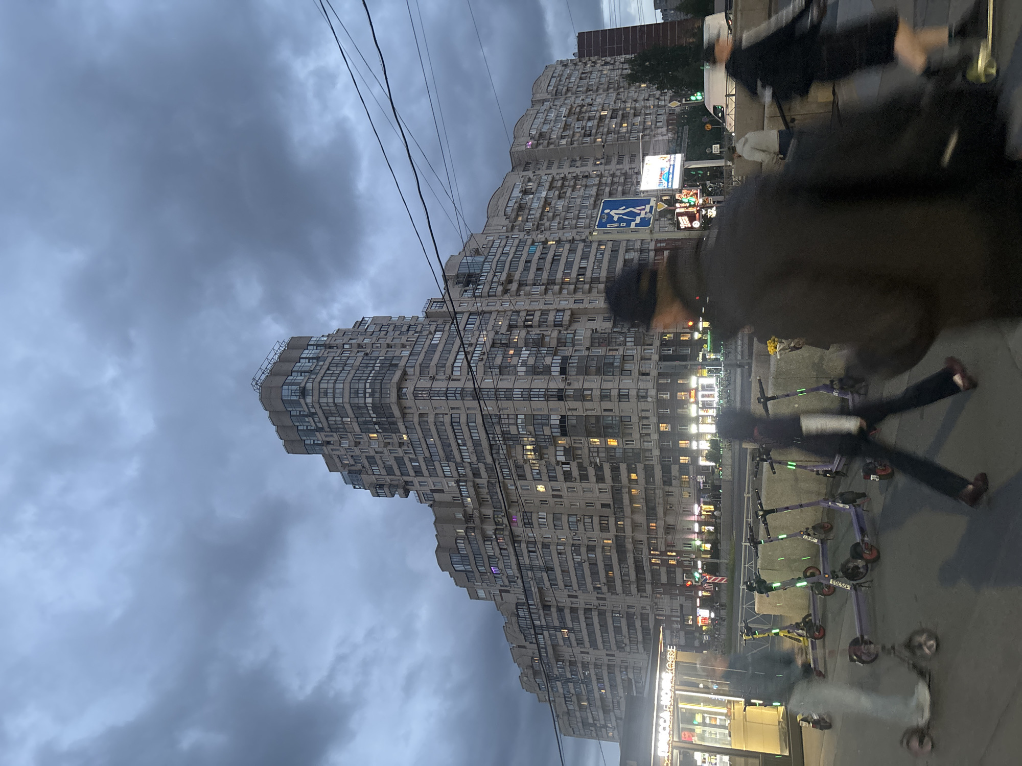 The image captures an evening street scene in Saint Petersburg, Russia. A high-rise apartment building dominates the background, its many windows lit up. Overcast skies indicate the possibility of rain. A row of electric scooters is parked on the right side of the frame, near the sidewalk. Passersby walk along the sidewalk, some blurred due to motion. Street lights and store signs illuminate the street level. A pedestrian crossing sign is visible near the building. Overhead power lines run across the sky. The scene is a typical urban landscape.