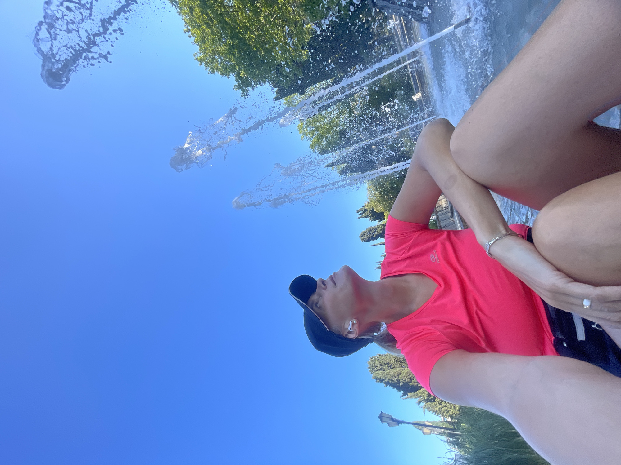 A low-angle, upright portrait captures an individual outdoors on a clear, sunny day in Sochi, Russia. The person, likely female, is positioned in the foreground, wearing a bright fuchsia-colored short-sleeved athletic t-shirt with the brand "KALENJI" faintly visible, a black baseball cap, and white wireless earbuds. A silver bracelet is on one wrist, and a ring is visible on a finger. Their head is tilted back, and they are looking upwards.
The background is dominated by a vibrant blue sky, against which multiple streams of water from a large fountain are actively spraying upwards. Green foliage from trees is visible, framing the scene and suggesting a park or public garden setting. A street lamppost stands in the background to the right. The bright light indicates it is daytime. The image conveys a sense of enjoying the pleasant outdoor environment and the fountain's display.
