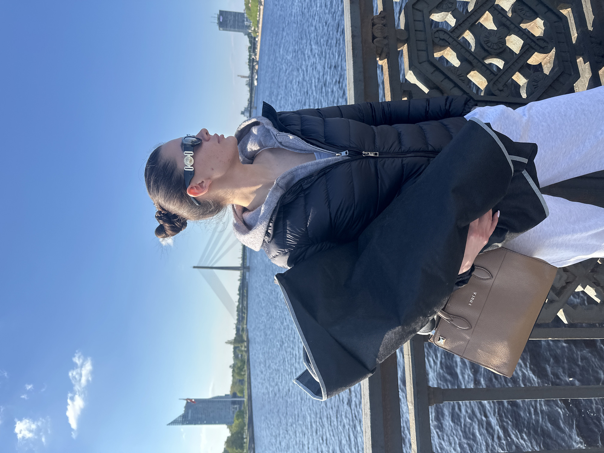 A young woman stands on the Vansu Bridge in Riga, Latvia, looking upward. She is wearing sunglasses, a black puffy jacket over a grey hoodie, and white pants. She is carrying a tan Furla handbag. In the background is a body of water with the city skyline visible, including the Latvian Academy of Sciences building and the Cable Bridge. The sky is blue with a few clouds. The metal railing of the bridge is visible in the foreground.
