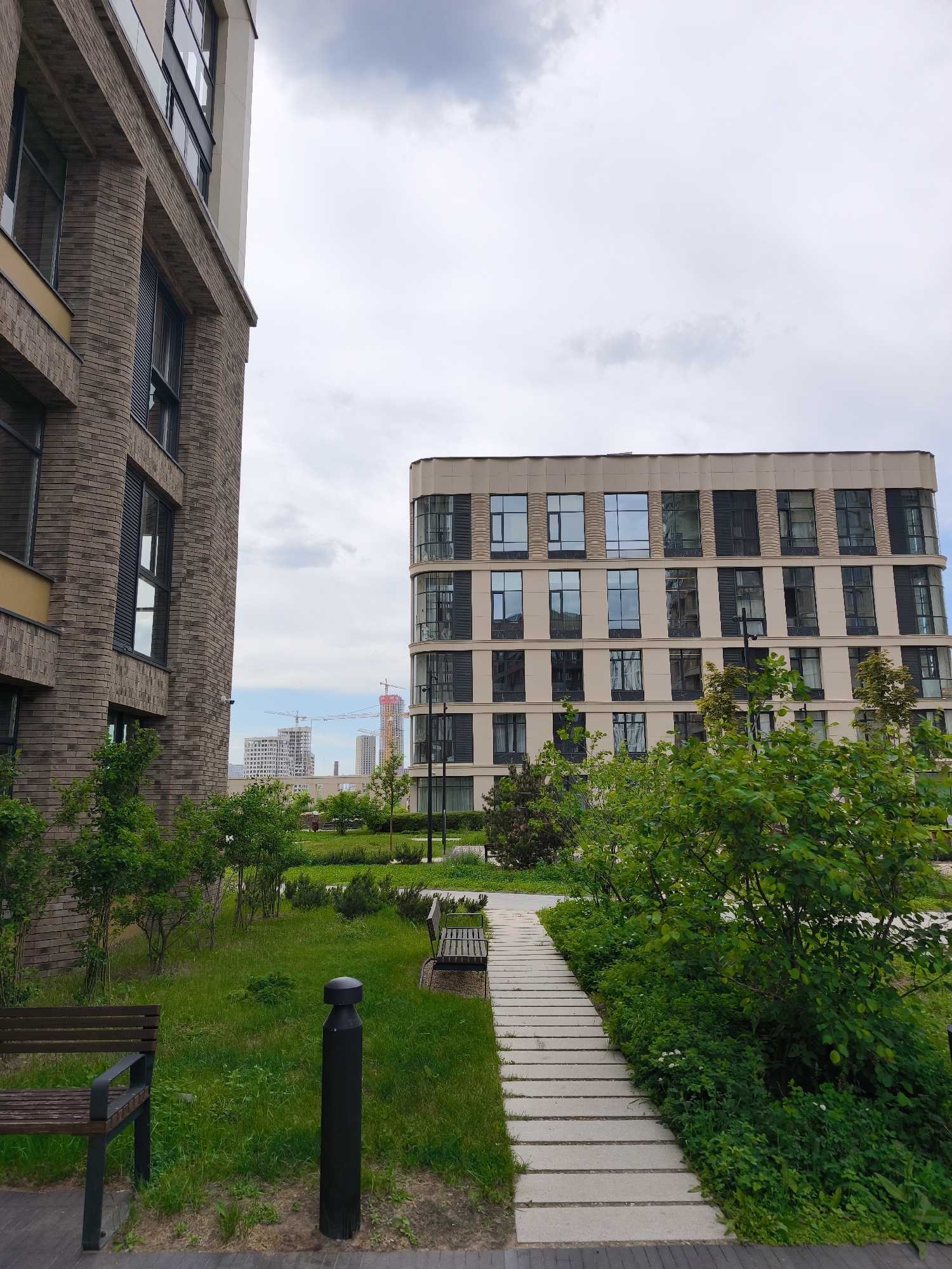 The image displays a modern urban landscape in Moscow, Russia, featuring a landscaped green space nestled between two contemporary multi-story buildings, all under an overcast sky.
On the left, a prominent building features a facade of dark brown brickwork, contrasted with lighter cream-colored sections and large, dark-framed windows. The building on the right, further in the scene, is characterized by a light beige exterior, large windows (some with a curved design), and dark vertical elements, giving it a sleek, modern appearance with a rounded corner.
A light-colored pathway, constructed from rectangular pavers, winds through the central green area. This space is richly planted with lush green grass, various bushes, and small trees. Two dark wooden benches with metal frames are visible, one in the foreground to the left and another further along the path, providing resting spots. Dark, cylindrical bollards, possibly light fixtures, are also interspersed within the greenery.
In the distance, beyond the immediate buildings and green space, several structures are under construction, indicated by the visible cranes towering against the cloudy sky, suggesting ongoing development in the urban environment. The sky itself is overcast with light grey clouds, indicating a cloudy daytime. No people or visible text are present in the scene, which focuses on the architectural design and natural elements of the modern complex.