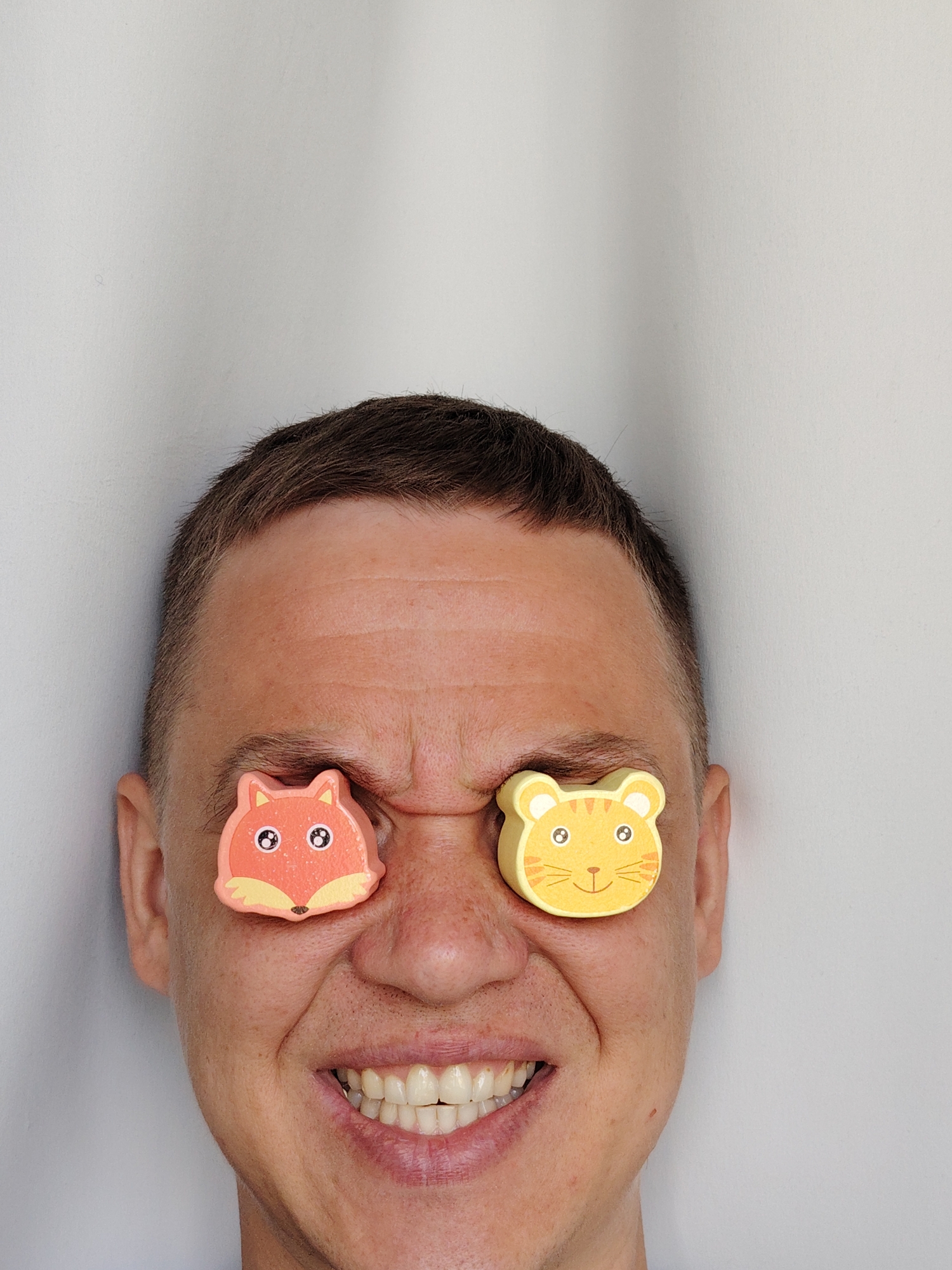 A Caucasian male with short brown hair is positioned centrally in the frame, smiling with clenched teeth. His eyes are obscured by small, flat wooden toys - a fox on the left and a tiger on the right. The background is a plain, light-colored wall. The image was taken in Chaikovsky, Russia.