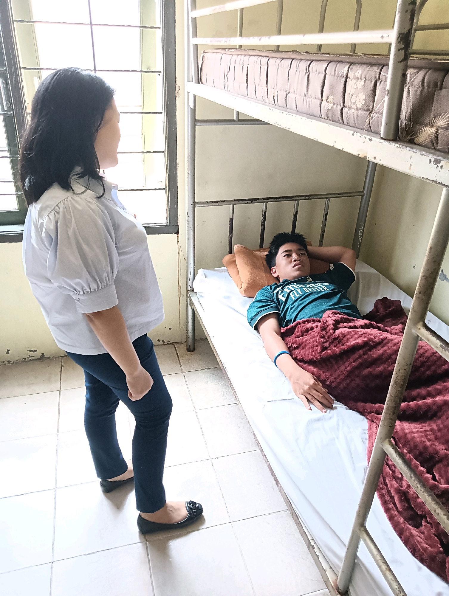 A woman stands facing a young man lying in a bunk bed in a room that appears to be a dormitory or a small dwelling. The woman, with dark hair and wearing a white collared shirt with puffed sleeves and dark pants, stands near a window with metal bars. The young man, wearing a teal t-shirt and a maroon blanket, is lying on his back in the lower bunk, with his head resting on a pillow. The room is sparsely furnished with a metal bunk bed and tiled flooring. The overall impression is a moment of quiet interaction or observation within a simple living space. The location is identified as Tomohon, Indonesia.