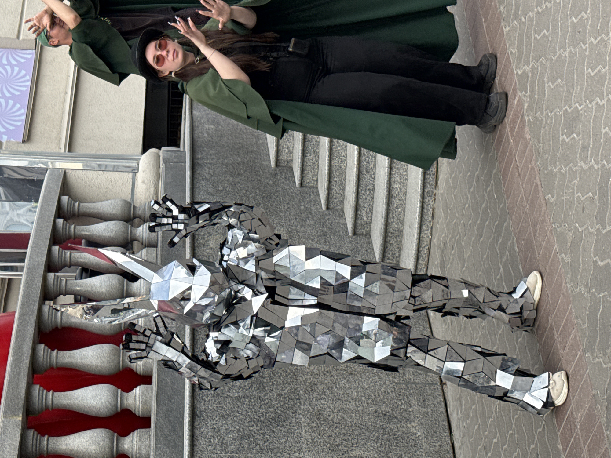 In Kazan, Russia, two individuals are captured in a vertical-oriented photograph. In the foreground, a person is dressed in a full-body costume made of mirrored, geometric panels, giving them a multifaceted, almost robotic appearance. This costume features a pointed headpiece, suggestive of an animal or creature, and covers the entirety of their body down to white sneakers. The mirrored surface reflects the surroundings, including the patterned pavement and parts of the building.
Behind the mirrored figure, two people are partially visible, wearing dark green capes. One person, wearing sunglasses and a black hat, is looking towards the camera with an open-mouthed expression, suggesting surprise or engagement. Their hands are raised. The other person, further back, also has their hands raised in a similar gesture. They are both wearing black pants and dark shoes.
The scene appears to be an outdoor urban setting, with a stone-tiled walkway and a building with a textured facade and ornate railings in the background. The lighting suggests it is daytime. The overall mood seems to be one of performance or street entertainment, with the mirrored costume likely being a highlight for onlookers. No visible text is present in the image.