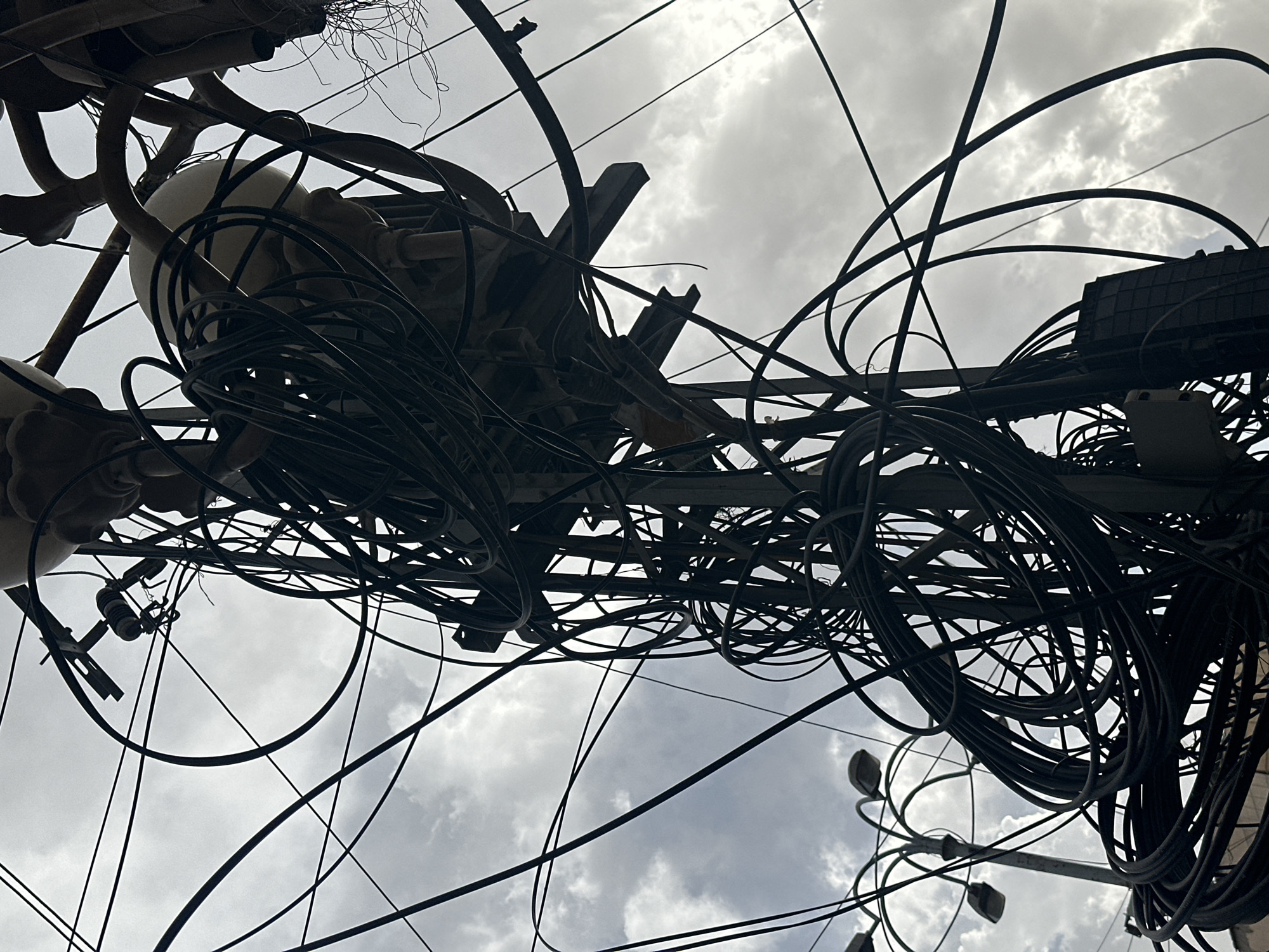 The image depicts a dense network of power and communication lines in Quetta City Tehsil, Pakistan. The tangle of black cables, suspended overhead, are attached to poles and equipment, obscuring the view of the sky with its overcast conditions. Street lights are visible among the cables.