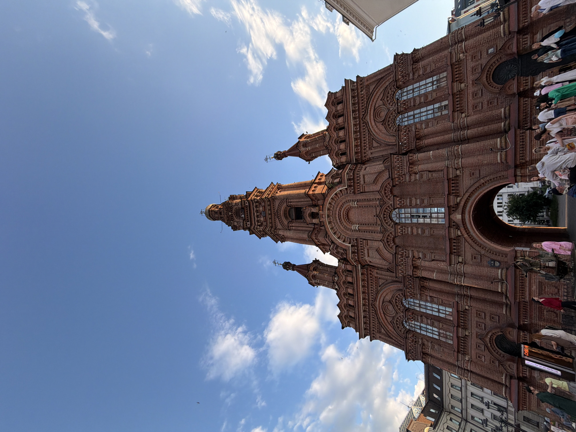 The image displays a low-angle, wide-angle view of the Epiphany Cathedral (Bogoyavlensky Cathedral) in Kazan, Russia, under a partially cloudy blue sky during what appears to be late afternoon or early evening.
The central subject is the massive brick cathedral, constructed predominantly from reddish-brown brick. Its architecture features a very tall, multi-tiered central bell tower topped with a cross, flanked by two smaller, similarly styled spires, each also surmounted by a cross. The facade is intricately detailed with numerous arched windows of varying sizes, decorative brickwork, recessed panels, and corbelling. A large, prominent archway pierces the base of the cathedral, allowing passage. Sunlight illuminates the upper sections and the right side of the structure, casting some subtle shadows.
At the base of the cathedral and in the foreground, approximately 20-30 individuals are present. They are dressed in casual clothing, including t-shirts, dresses, and light pants, and are engaged in various activities such as walking, standing, and conversing in small groups. On the left side, near an entrance, a sign with Cyrillic text, partially visible as "СУВЕН...", is affixed. To the right of the central archway, a dark, sculptural element, possibly part of a fountain, is visible near a group of people.
The sky is bright blue with scattered white, wispy clouds, more concentrated on the left side. The lighting suggests a clear day. Portions of other buildings are visible on the periphery: a multi-story building with light-colored walls and dark roofs on the far left, and a white architectural detail with a dark gutter on the far right. Faint, dark specks, possibly birds, are discernible in the sky.