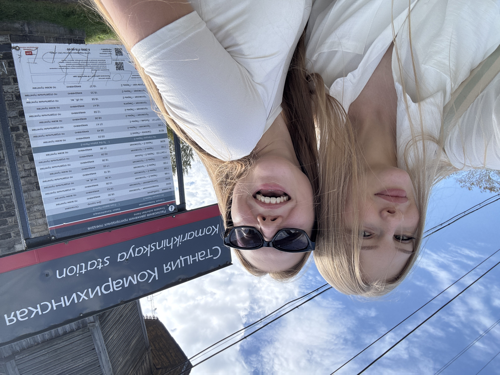 The image shows two young women posing for a selfie outdoors in front of a train station sign. The sign reads "Станция Комарихинская Komarikhinskaya station" and displays a timetable for suburban trains. The women are smiling and looking at the camera. The woman on the left has blonde hair and is wearing a white shirt. The woman on the right has blonde hair and is wearing sunglasses and a white t-shirt. The background shows a blue sky with some clouds and overhead wires. The train station appears to be in a rural or suburban area, possibly in Russia, given the Cyrillic text on the sign. The overall impression is a casual and happy moment captured on camera.