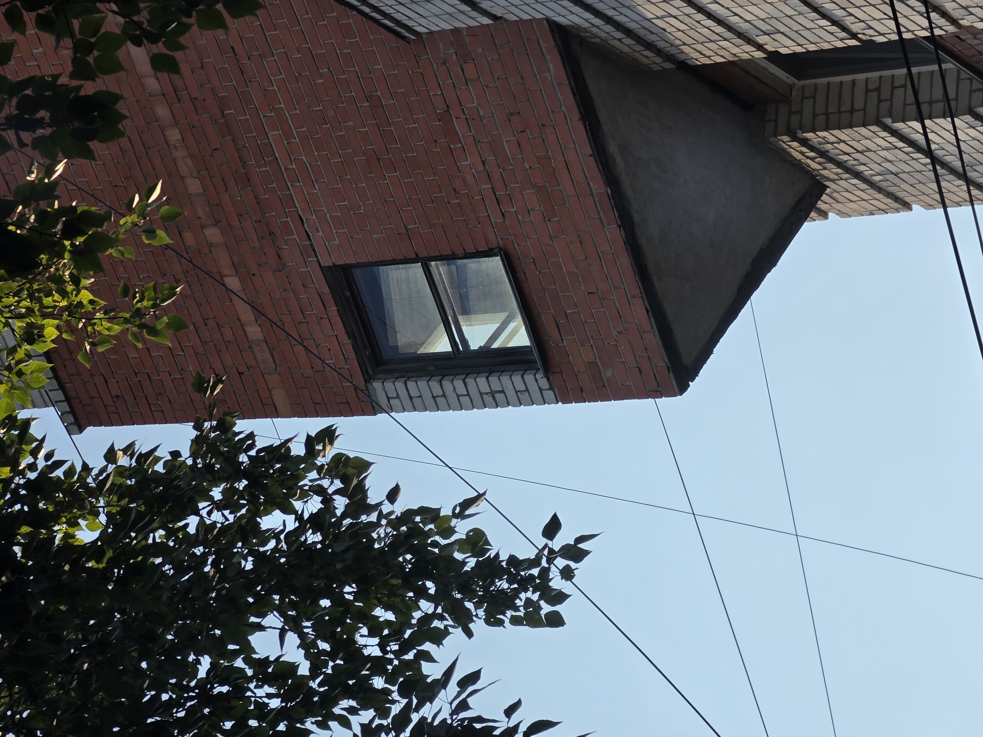 An exterior, eye-level shot of a residential building in Nizhny Novgorod, Russia shows a section of a brick wall protruding at an odd angle. The wall is constructed from red brick, with a single window set into it. Below the window, a concrete overhang extends outward, creating a sharp triangular shape. To the left of the protruding section, a portion of a building clad in lighter colored brick is visible. The sky is clear and blue, with several dark wires crisscrossing the frame. Lush green tree branches with dappled sunlight are hanging from the top left corner of the image. The angle and architecture suggest this may be a unique or older section of the building, possibly indicative of Soviet-era construction methods.