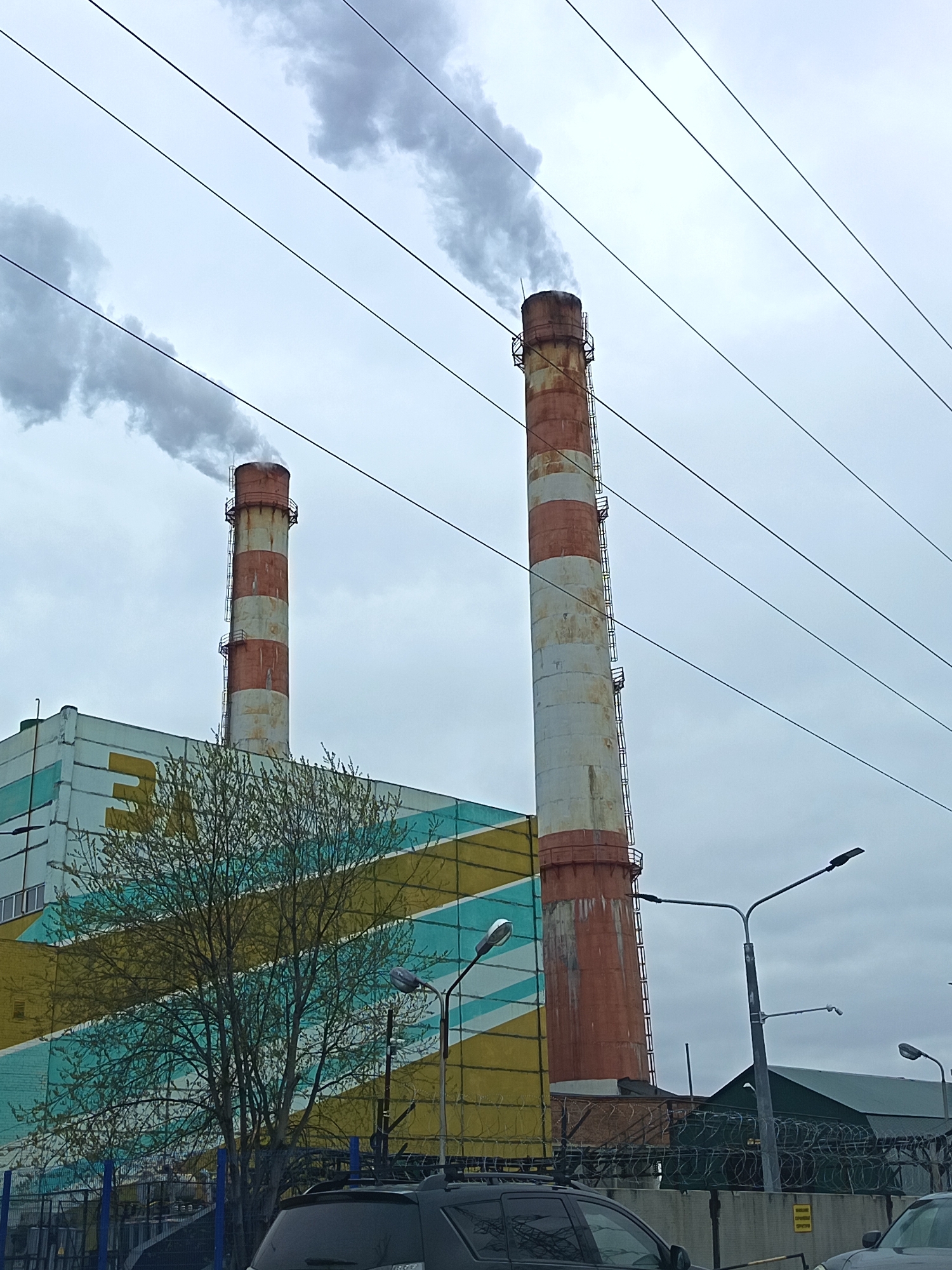 The image captures a daytime view of an industrial area in Nizhnevartovsk, Russia, characterized by a power plant emitting smoke from two tall, striped chimneys. The chimneys, alternately colored in white and orange-red bands, stand behind a large, rectangular building with horizontal stripes of yellow and teal. A leafless tree obscures part of the building's facade. Overhead, a network of power lines stretches across the overcast sky. Streetlights are visible along the perimeter, and a barbed-wire fence secures the area. Several vehicles are parked in the foreground, including a black SUV with a roof rack. The overall impression is one of a typical industrial landscape in an urban setting.