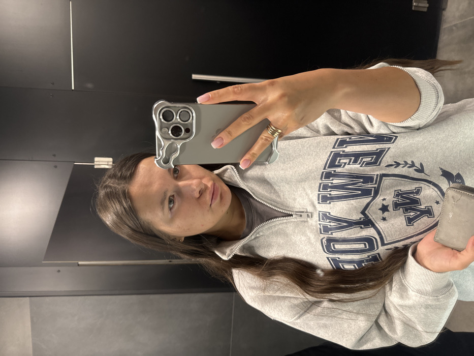 The image displays a mirror selfie taken by a young person, likely female, indoors. The scene is rotated 90 degrees clockwise, suggesting it was captured with the phone held horizontally and then uploaded.
The person has long, dark brown hair that falls over their shoulders and chest. They are wearing a light gray pullover or sweatshirt with a half-zip neckline. The sweatshirt features a prominent dark blue graphic print on the front. Above a shield or crest design, the word "NEW" is clearly visible, followed by letters that appear to spell "WEIL" or "WELL". Inside the crest, the text "ROY" is legible, and below it, two stars contain the letters "Y" and "M" respectively.
The person's face is visible, looking towards the camera (phone). They have dark eyes and a neutral expression. Their right hand holds a smartphone, which is dark gray or silver-colored and encased in a distinctive clear or metallic-looking case with wavy, "melted" edges, particularly around the triple camera module. The person's nails have a French manicure style with pink bases and white tips. Rings, possibly three simple bands, are visible on a finger of their right hand. In their left hand, they hold a small, light beige or gray wallet or cardholder, which appears somewhat worn.
The background, visible in the mirror reflection, consists of dark-colored panels or walls, possibly black or dark gray, with visible seams or joints creating horizontal lines. A silver-colored handle or bar is also reflected on the dark surface. The setting appears to be a functional indoor space, such as a restroom or changing area.