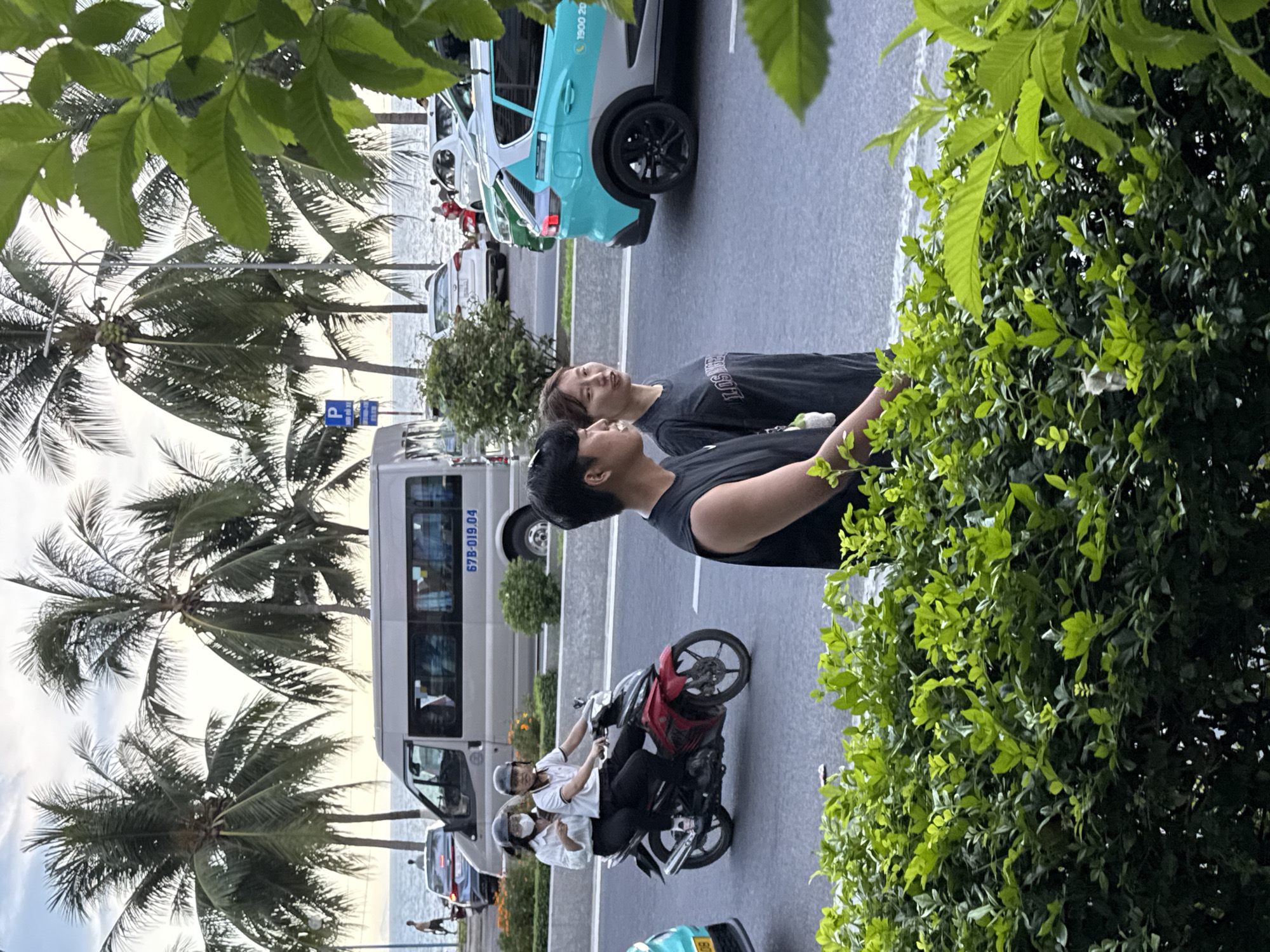 The image depicts a street scene in Nha Trang Ward, Vietnam, with a focus on a motorcycle carrying two people and a bus. The sun is setting, casting a warm glow over the scene, and the weather appears to be clear and pleasant.
In the foreground, a motorcycle is the central object. It is red and black and has two individuals riding on it. The person in the front is wearing a white shirt and a helmet, while the person behind them is wearing a white shirt and a face mask. They are both looking forward, seemingly engaged in conversation.
Behind the motorcycle, a white bus is parked on the side of the road. The bus has a license plate that reads "67B-019.04". Further in the background, palm trees line the street, suggesting a tropical or subtropical location.
To the right of the image, lush green foliage obscures part of the scene, adding a natural element to the urban setting. Two individuals, a man and a woman, are standing and looking to their left. The woman is wearing a black t-shirt with the word "LOS" visible, and the man is wearing a black t-shirt. Their expressions are not clearly discernible, but they seem to be observing something off-camera.
In the background, a turquoise and white car is parked, with the text "1900 202" visible on its side. Another car, a white SUV, is also parked on the street. The presence of multiple vehicles suggests a moderately busy street. The ocean is visible in the distance, with calm waters, indicating the proximity to the coast.