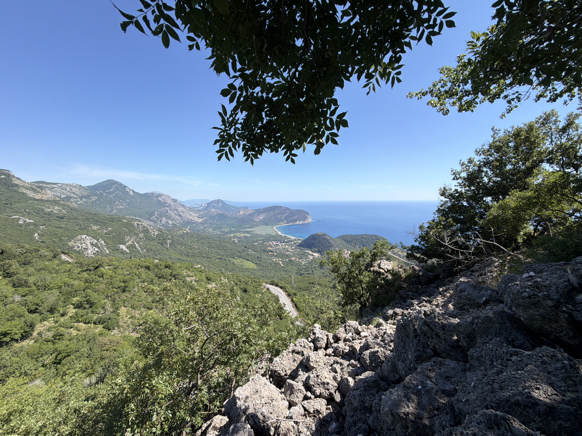 This is a panoramic, eye-level, outdoor photograph taken from a rocky, elevated vantage point. The scene overlooks a coastal landscape with a bright blue sky. In the foreground, large, rough-textured gray rocks dominate the lower right portion of the image. Lush green trees and foliage are visible in the foreground and middle ground, framing the expansive view.
The middle ground features a winding asphalt road leading down a green, forested mountainside. Further in the distance, the landscape transitions to a bay with a sandy beach, dotted with small buildings that form a village. Beyond the village, a peninsula juts into the calm, deep blue sea. The horizon line is sharp, with distant hills and mountains visible on either side of the bay. The overall impression is one of natural beauty and a serene coastal environment.
The location is identified as being in Novoselje, Montenegro.