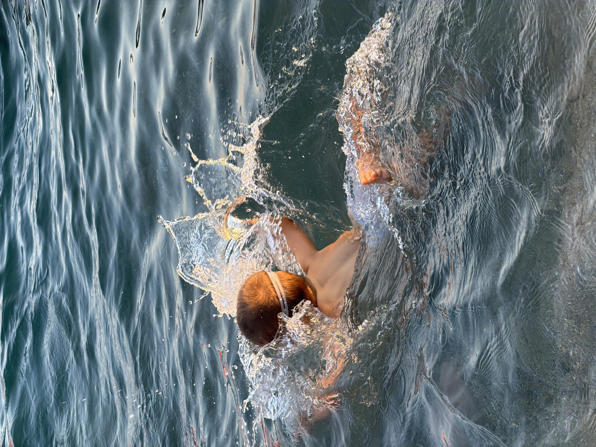 A young boy with goggles is swimming in the sea at Bečići, Montenegro. He is splashing water with his arms and legs, creating a large spray around him. The water is a deep blue-green color, and the sunlight is reflecting off the waves. The boy is wearing white swim trunks. The scene appears to be a warm, sunny day.