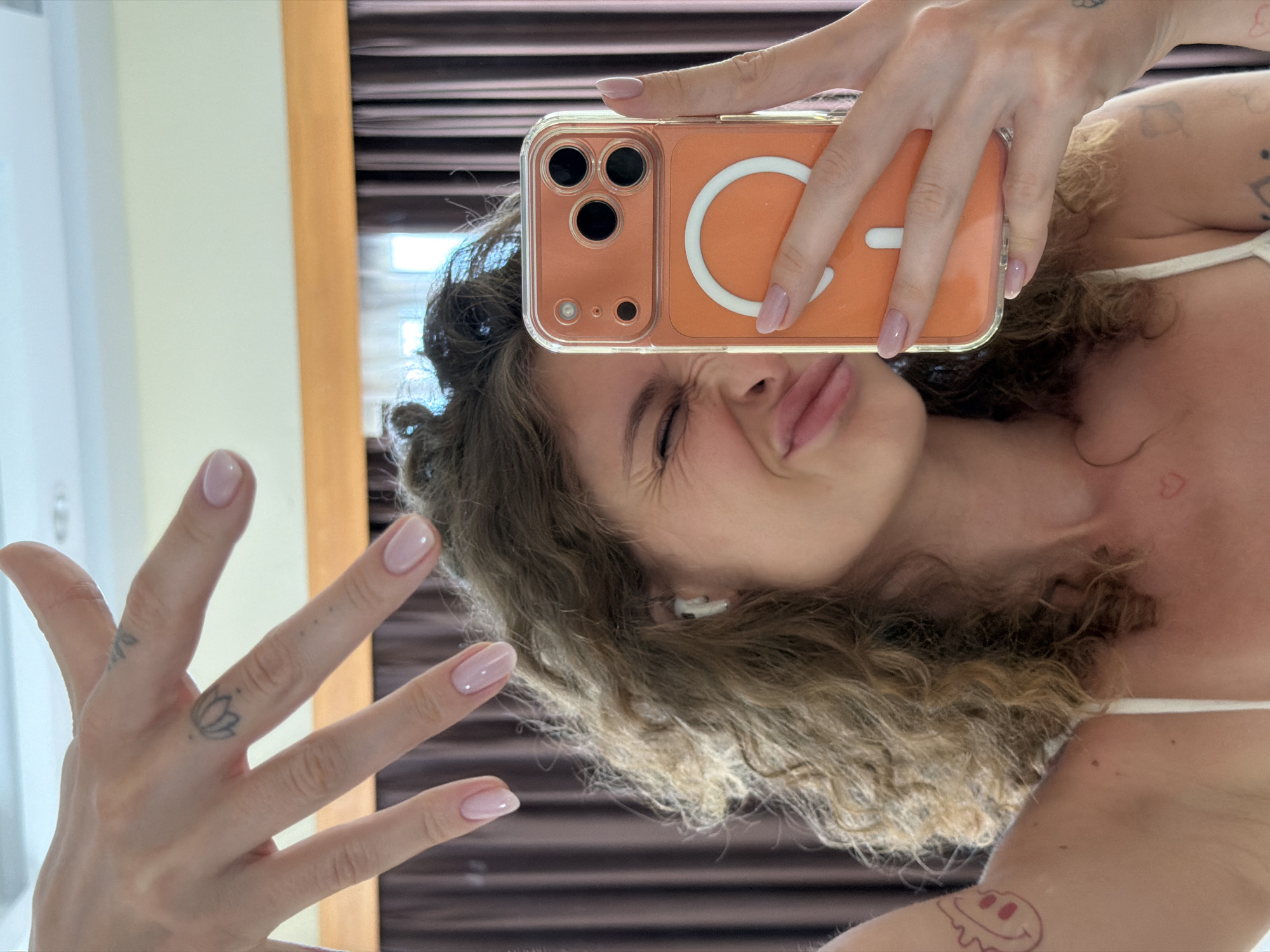 A light-skinned woman is taking a selfie in a mirror. She is wearing a light-colored top and has curly brown hair. She is holding an orange iPhone with a clear case, and making a pouting face at the camera. Her left hand is also visible, with a lotus flower tattoo on her middle finger. She has neatly manicured nails with a nude pink color. She has a tattoo of a heart on her collarbone and a melting smiley face tattoo on her arm. A white wireless earbud is visible in her ear. The background shows part of a cream-colored wall, a narrow brown wall, and blinds on a window. The selfie appears to be taken indoors. It is unknown if this picture was taken in North Kuta, Indonesia.