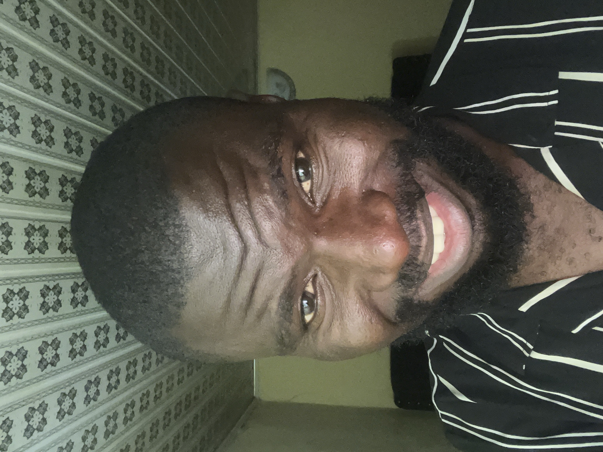 A close-up, indoor shot shows a man with dark skin, a close-cut haircut, and a thick beard, wearing a black and white striped shirt and smiling. Behind him is a decorative wall and a section of a green wall. A white object is affixed to the green wall. The location is in Gura Top, Nigeria.