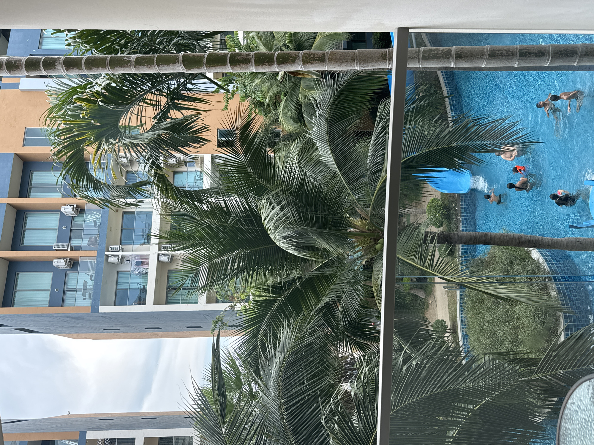 The image captures an outdoor scene from an elevated perspective, likely a balcony, overlooking a hotel or apartment complex pool area. In the foreground, numerous lush green palm trees with prominent trunks and fronds dominate the view, some bearing visible coconuts. A white railing or balcony structure is partially visible in the bottom right corner, suggesting the vantage point.
Below the dense foliage of the palm trees, a vibrant blue swimming pool with tiled patterns is visible. Several people, including adults and possibly older children, are enjoying the water. Approximately six to eight individuals are present in the pool; some are standing, while others appear to be swimming. One person is wearing an orange life vest, and another is in what looks like a black wetsuit. A bright blue water slide empties into the pool on the left side. Surrounding the pool are some green shrubs and a paved area.
In the mid-ground and background, multi-story buildings with modern architecture are seen. These structures feature balconies with outdoor air conditioning units and large windows. The buildings are painted in alternating sections of muted orange/beige and shades of blue or grey. Above the buildings, a clear, light blue sky with scattered clouds suggests fair weather. The scene appears to be set during daytime, likely under bright sunlight given the clear visibility and shadows. The overall atmosphere is one of leisure and relaxation. There is no discernible text visible in the image.