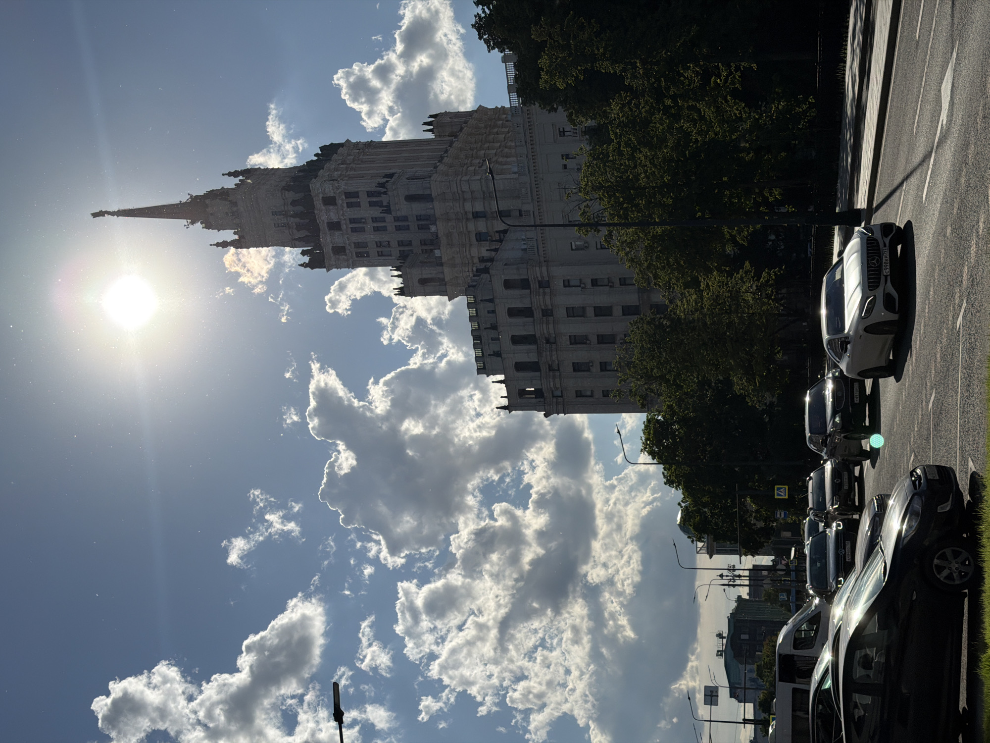 The image captures a vibrant street scene in Moscow, Russia, featuring a prominent Stalinist Empire style skyscraper under bright sunshine. The tall, ornate building, characteristic of Moscow's "Seven Sisters," stands majestically with its intricate tiers and a distinctive spire reaching towards a blue sky.
The sun, positioned to the left of the building, casts a bright lens flare and illuminates parts of the facade, creating strong contrasts and shadows. The sky is partly cloudy with fluffy white cumulus clouds.
In the foreground and to the right, a street runs parallel to the building, lined with parked cars. A white sedan is prominently visible, and its license plate "c998xp 777" is clearly legible, with "777" being a regional code for Moscow. Further down the street, other vehicles are parked, and a yellow pedestrian crossing sign is discernible. Lush green trees line the street, partially obscuring the lower parts of the skyscraper.
The time of day appears to be either early morning or late afternoon, given the angle of the sun and the long shadows cast. The weather is clear and sunny. The scene depicts a typical urban environment, blending historical architecture with the everyday life of a modern city.