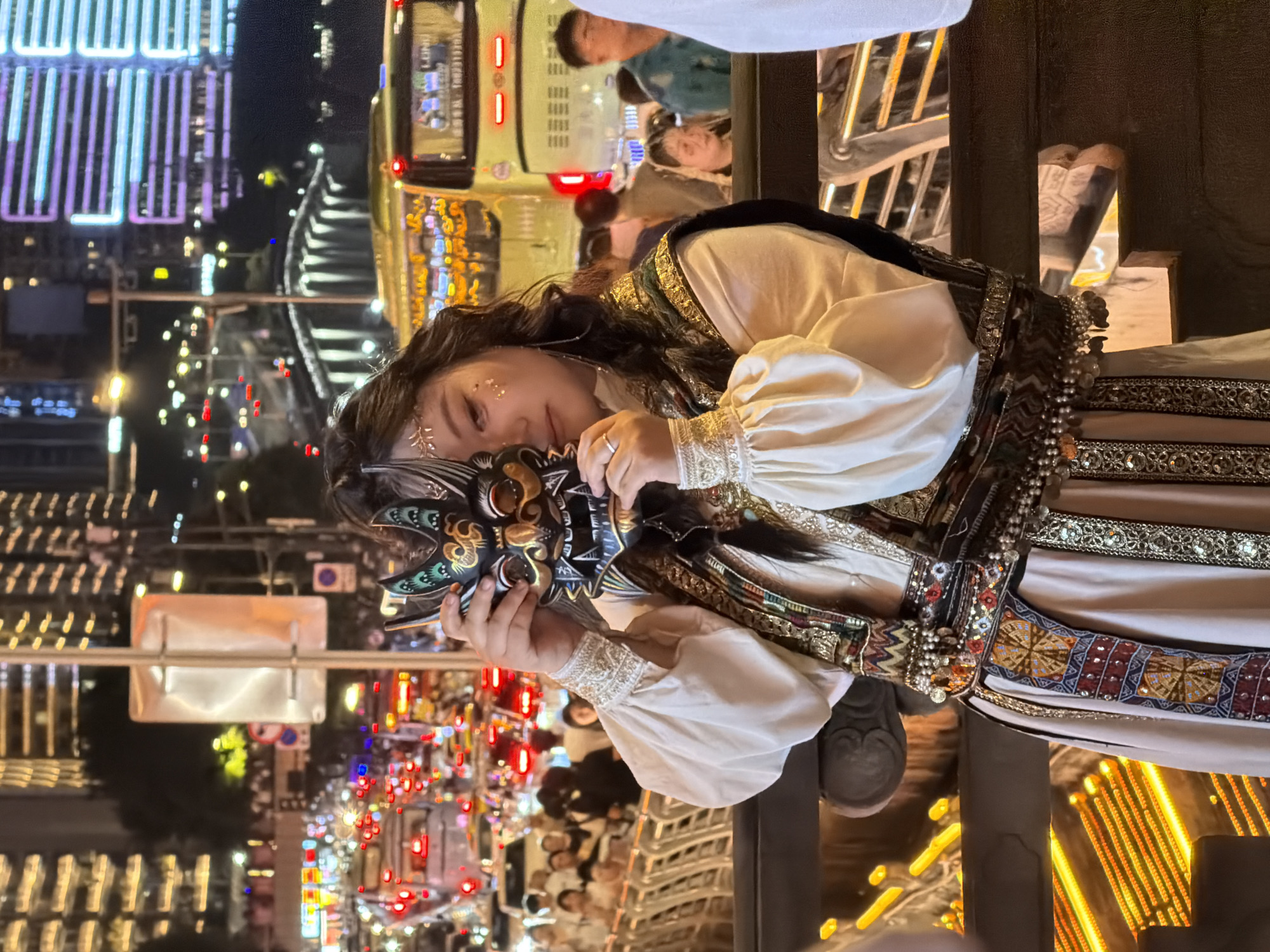 In Yuzhong District, China, a young woman stands outdoors at night, holding a mask in front of her face. She is wearing a traditional-style outfit with a white, long-sleeved blouse, a richly decorated vest, and a sash. Her hair is dark and long, and she has glitter applied to her forehead. Behind her, the scene is a bustling city street at night, filled with traffic, illuminated buildings, and other pedestrians. The urban environment includes a large yellow bus on the right and a bridge in the background. The overall impression is of a vibrant, lively urban setting with a focus on the woman and her mask, suggesting a cultural or festive event.