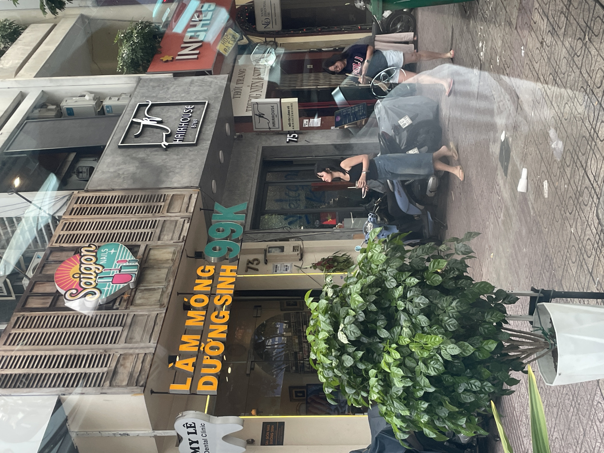 In Thủ Đức City, Vietnam, a street scene unfolds, captured through a reflective surface. A white potted plant with lush green foliage stands prominently in the foreground, its base slightly obscured by the reflection. Behind it, a vibrant sign for "Saigon Nails" is visible, featuring a sunburst motif and images of nail polish bottles. Adjacent to this is another sign, "LÂM MÔNG DƯỜNG SINH 99K," illuminated in bright orange and green, indicating a service likely related to nail care or beauty.
Further down the street, a sign for "HAIRHOUSE" is mounted on a gray facade. Across the street, a red sign with white lettering spelling "INCHES" is partially visible. The pavement is paved with square tiles, and various motorbikes are parked along the sidewalk, some covered with protective sheets.
Two women are present in the midground. One, wearing a black tank top and denim skirt, stands holding a drink with a straw, looking to her left. The other, in a navy t-shirt and white shorts, is to her right, also holding a drink and appears to be looking down. Reflections on the surface through which the photo is taken add a layer of abstraction to the scene, obscuring details and distorting shapes. The overall impression is a busy urban environment, possibly a commercial street, with a focus on beauty and personal care services.