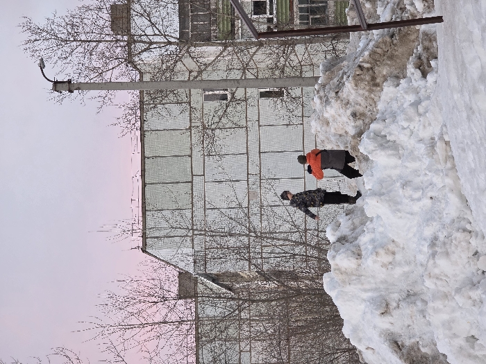 The image depicts an outdoor winter scene in the city of Chaikovsky, Russia, characterized by significant snow accumulation and bare deciduous trees under a pale pink and grey sky. In the foreground, large, uneven piles of snow, some appearing dirty with brown discoloration, dominate the lower half of the frame. Two individuals are positioned on these snow piles. The person on the left stands upright, dressed in a camouflage-patterned jacket, dark pants, and dark boots, with a dark hat or hood. The person on the right is crouched or bent over, wearing an orange and grey jacket, dark pants, and a dark hat, holding a small dark object, possibly a camera or controller, pointed upwards. In the background, a multi-story residential building with a light-colored, gridded facade is visible, partially obscured by the bare trees. The building exhibits some dark-framed windows and a section of green paneling on its right side. A grey utility pole with a streetlamp fixture extends vertically on the right side of the image, positioned in front of the building.