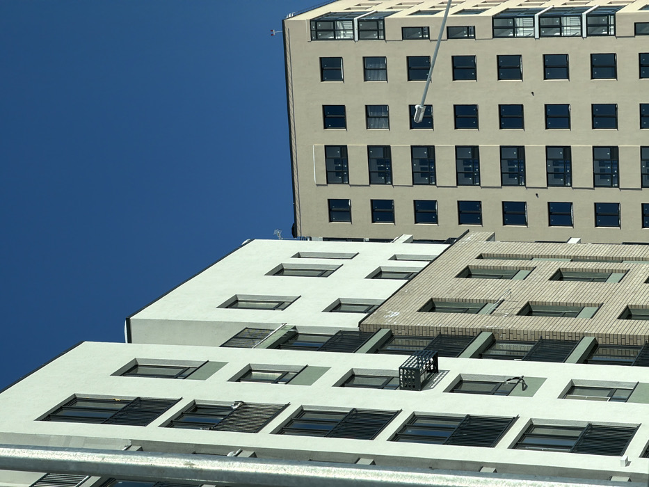 The image captures a detailed view of modern, multi-story residential buildings under a clear blue sky in Tyumen, Russia. The foreground features a light-colored building, primarily white with subtle light green accents between window sections, equipped with dark-framed windows and black metal balconies. A lower portion of this structure transitions to a light beige brick facade. A small, open metal platform-like structure protrudes from one floor on the white building. To the right, a taller, uniform light tan/beige building is visible, characterized by numerous dark-framed, rectangular windows and a section of glazed, enclosed balconies on its rightmost edge. Traces of snow are visible on some window sills and flat rooftop surfaces, indicating cold weather conditions. A white street light fixture on a pole is positioned in the mid-ground, partially obscuring the beige building. No individuals or vehicles are present in the visible scene.