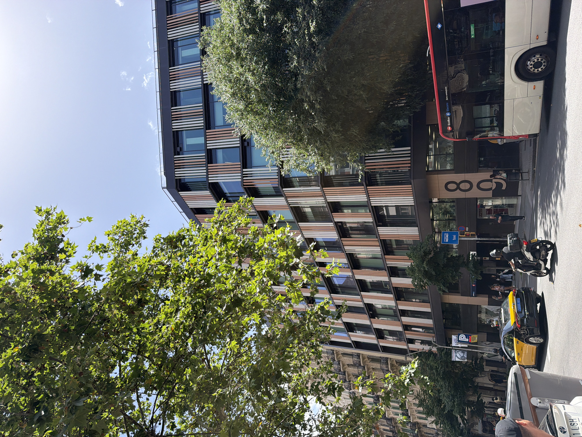 This is a medium-shot, eye-level outdoor photograph taken on a sunny day in Barcelona, Spain, likely on Avinguda Diagonal, given the architecture and street elements. The scene captures a street view with modern and older buildings, vehicles, and pedestrians.
In the foreground, on the right, is the rear portion of a white bus with a red stripe along the top. The bus has "EcoBus Electric Zero emissions" written on its side. To the left of the bus is a dark-colored building with a large, stylized "808" sign on its facade. In front of this building, a few pedestrians are visible, including a person standing near a trash receptacle and others walking away from the viewer.
In the middle ground, a yellow and black taxi is stopped in the street. A person is seated in the driver's seat. To the right of the taxi, a person is riding a white scooter with a black helmet. Further to the left, a grey car with a partial license plate "1 LSH" is visible from the side.
The background features a prominent, modern building with a facade composed of vertical slats in various shades of beige, brown, and grey, interspersed with windows. To the left of this building is an older, cream-colored building with balconies and ornate architectural details, indicative of early 20th-century construction.
Large trees with green leaves are positioned in the left and right portions of the frame, partially obscuring the buildings. The sky is blue with scattered white clouds. Street signs are visible, including a blue sign with a white "taxi" symbol and a P parking sign.