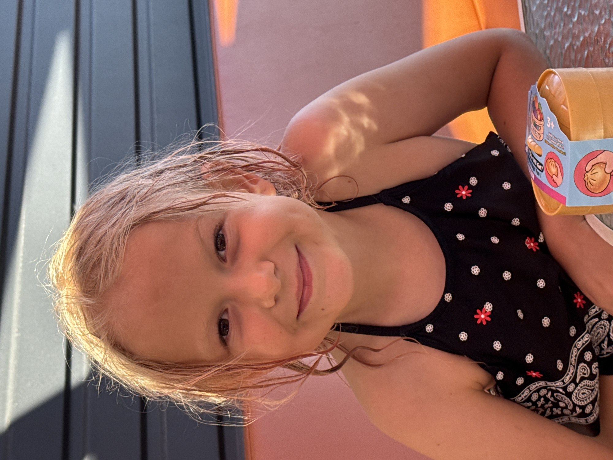 The image shows a young girl with light-colored, wet hair, smiling. She is wearing a black swimsuit with a pattern of white and red flowers. The girl is lying down, with her head tilted and looking towards the camera. In the background, there is a blurry orange object, possibly a toy bucket, and a pink surface. The scene appears to be outdoors, with dappled sunlight creating shadows on the girl's arm and on a dark, slatted surface to the left. The lighting suggests it might be daytime, possibly late afternoon due to the warm tone of the light. The girl's expression is happy and relaxed. There is text visible on the orange object, which appears to be a toy container. The text includes "3+" and images of food items, indicating it is a children's toy. The overall impression is a casual, summery moment captured outdoors.