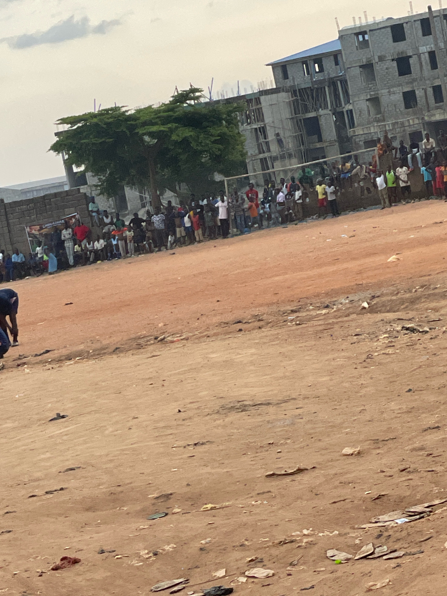 The image depicts a dusty, open field with scattered debris, likely in Abuja, Nigeria, given the context of similar construction and community scenes. In the background, a multi-story building is under construction, characterized by exposed concrete blocks and scaffolding. A large tree with dense green foliage stands near the construction site.
A crowd of people, predominantly young males, are gathered along a low wall or barrier in front of the construction. They appear to be spectators, observing something taking place in the foreground. Their attire suggests a casual, everyday setting.
In the foreground, a person, partially visible and wearing a dark blue shirt and dark pants, is bent over, appearing to be tending to something on the ground or perhaps engaging in a physical activity. The ground is a reddish-brown, dusty earth, typical of arid or semi-arid environments. The overall scene suggests a community gathering or sporting event taking place in an undeveloped area adjacent to a construction project. The sky is overcast, indicating a diffused light condition.