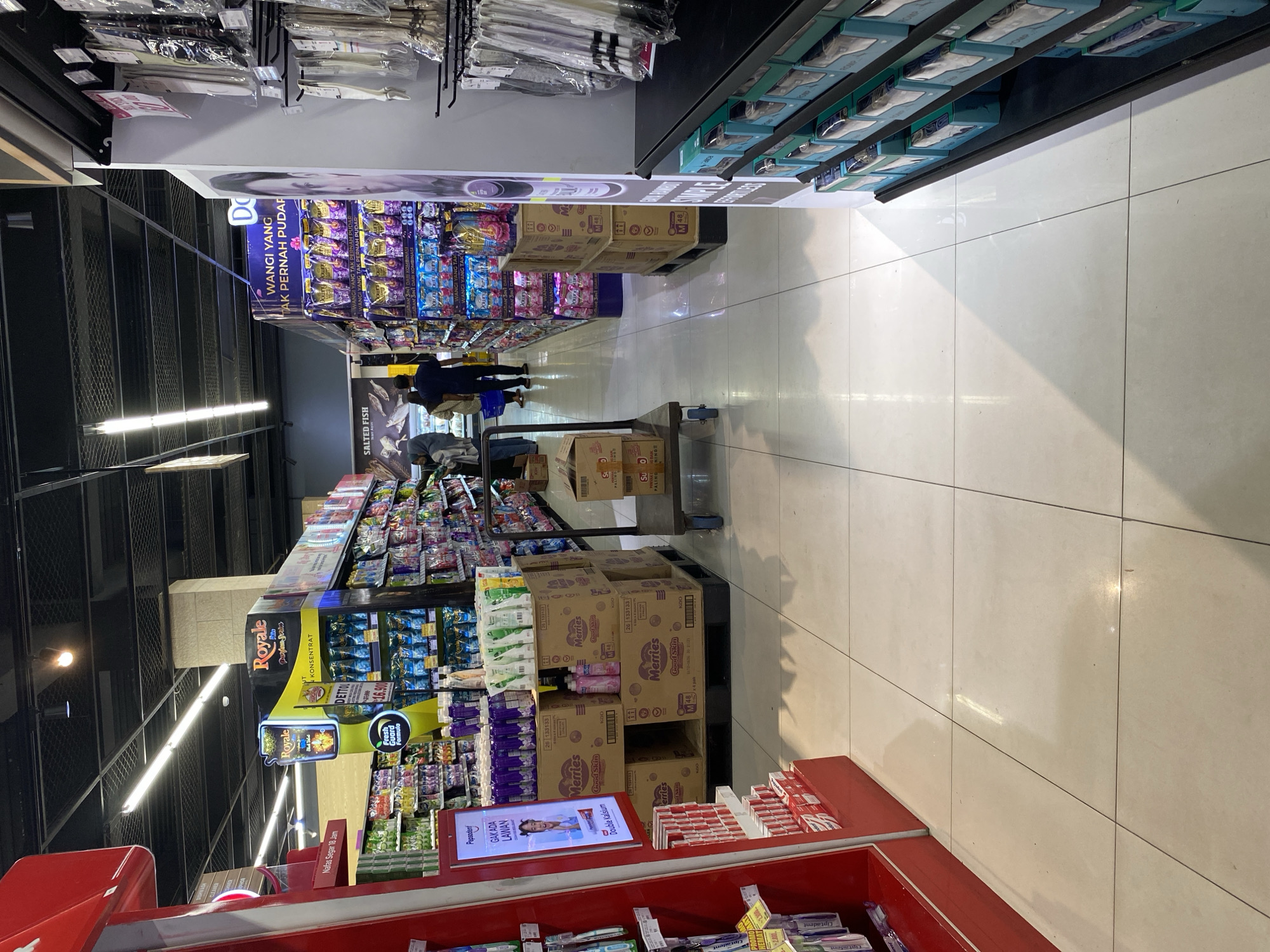 The image depicts an aisle in a supermarket in Banda Aceh, Indonesia. The aisle is lined with shelves stocked with various consumer goods. On the left, a red display unit features toothbrushes. In the center, shelves are filled with laundry detergent and other cleaning products, with boxes of diapers stacked on the floor. A shopping cart is positioned in the aisle. In the background, more shelves of products are visible, and two individuals are walking down the aisle. The floor is tiled, and fluorescent lighting illuminates the scene.