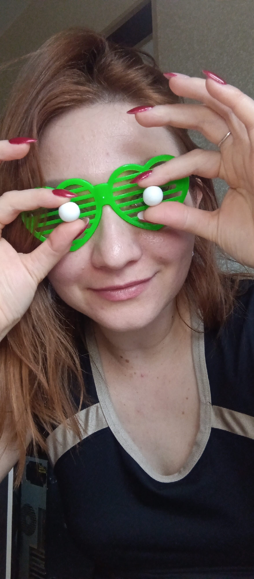 A close-up, eye-level shot features a person, likely female, with fair skin and long, reddish-brown hair, looking directly at the camera with a gentle, cheerful smile. She is wearing a pair of bright green, heart-shaped shutter shades, which cover her eyes. Playfully, she is holding two small, spherical white objects with her fingers, positioning them to appear as her eyes through the slats of the shades. Her hands are prominent, showcasing long, pointed fingernails painted in a vibrant red. A simple silver ring is visible on the ring finger of her right hand.
She is dressed in a dark-colored (possibly black or navy blue) top with a light-colored (beige or cream) trim around the v-neckline and shoulders. A few moles and freckles are visible on her neck and upper chest.
The background is indistinct, suggesting an indoor setting. A light-colored surface is behind her head, and a darker, textured area is to her right. On the far left, partially visible behind her shoulder, is what appears to be a grey computer tower or similar electronic equipment. The image conveys a lighthearted and playful mood. There is no visible text.