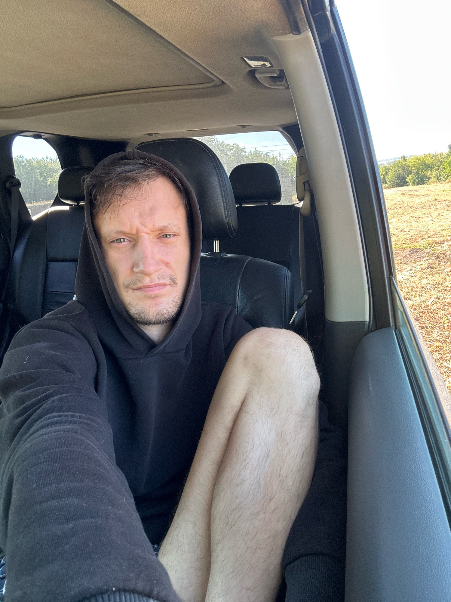 A man with short brown hair and a light beard, wearing a black hooded sweatshirt, is seated in the passenger seat of a car. The car's interior is visible, including black leather seats and a sunroof. The man is looking at the camera with a serious expression. His right leg is crossed over his left. Through the car windows, an outdoor environment with dry grass, trees, and a bright sky can be seen. The location is identified as Makiv, Ukraine.