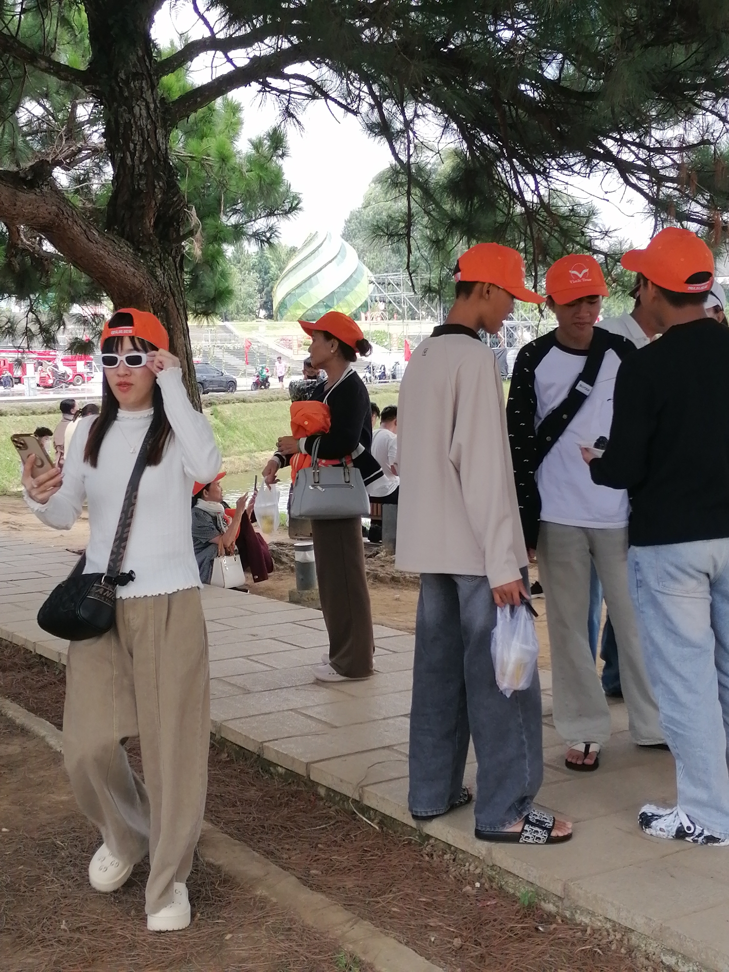 The image depicts a group of people gathered outdoors, likely in a park or public space in Da Lat, Vietnam. A young woman in the foreground, wearing a white long-sleeved shirt, khaki pants, white sunglasses, and an orange baseball cap, is taking a selfie with her smartphone. She has a black crossbody bag slung over her shoulder. Behind her, several other individuals are also present, some wearing orange baseball caps. The environment includes a paved walkway, dry ground with pine needles, and large trees with pine branches overhead. In the background, a distinctive spiraling green and white structure is visible, possibly a landmark or part of a tourist attraction. Red vehicles and other people can be seen further in the distance, suggesting a populated area. The overall atmosphere appears casual, with the group seemingly enjoying an outing.