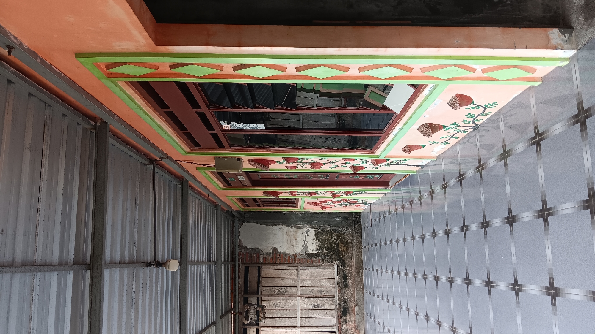 The image depicts the exterior of a building with an orange facade, located in the city of Sonobekel, Indonesia. The building features several windows with dark red frames and a patterned design along the exterior walls, consisting of green diamond shapes and red floral motifs. To the left, there is a covered walkway with a metal roof supported by beams. A light fixture hangs from the ceiling of the walkway. The ground is covered with square tiles arranged in a grid pattern. Further to the left, a section of the building reveals exposed brickwork and a wooden shuttered window, with a birdcage hanging above it. The scene is well-lit, suggesting it was captured during daylight hours.