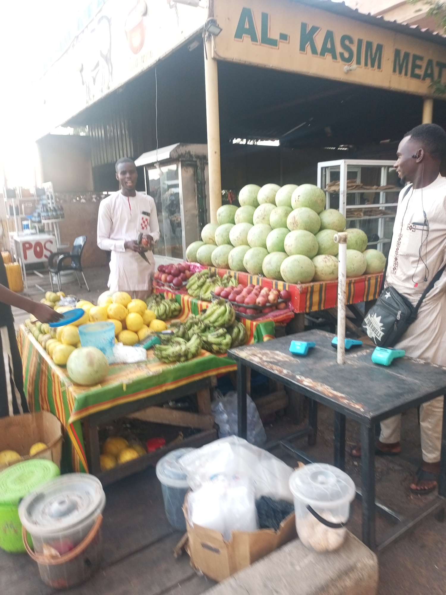 The image shows an outdoor fruit stand in Gombe, Nigeria, under a sign reading "AL-KASIM MEAT." A man in a light-colored traditional garment stands behind the stand, interacting with a customer who has a blue plate. The stand displays various fruits: melons piled high on one section, and lemons, plums, and bananas on another table covered with a striped cloth. Under the tables, there are buckets filled with fruits and some trash. A second man, also in a light-colored garment, is standing to the right, with a small black bag over his shoulder and earphones. A steel-framed table holds several small, turquoise-colored point-of-sale devices. In the background, there's a POS sign.
