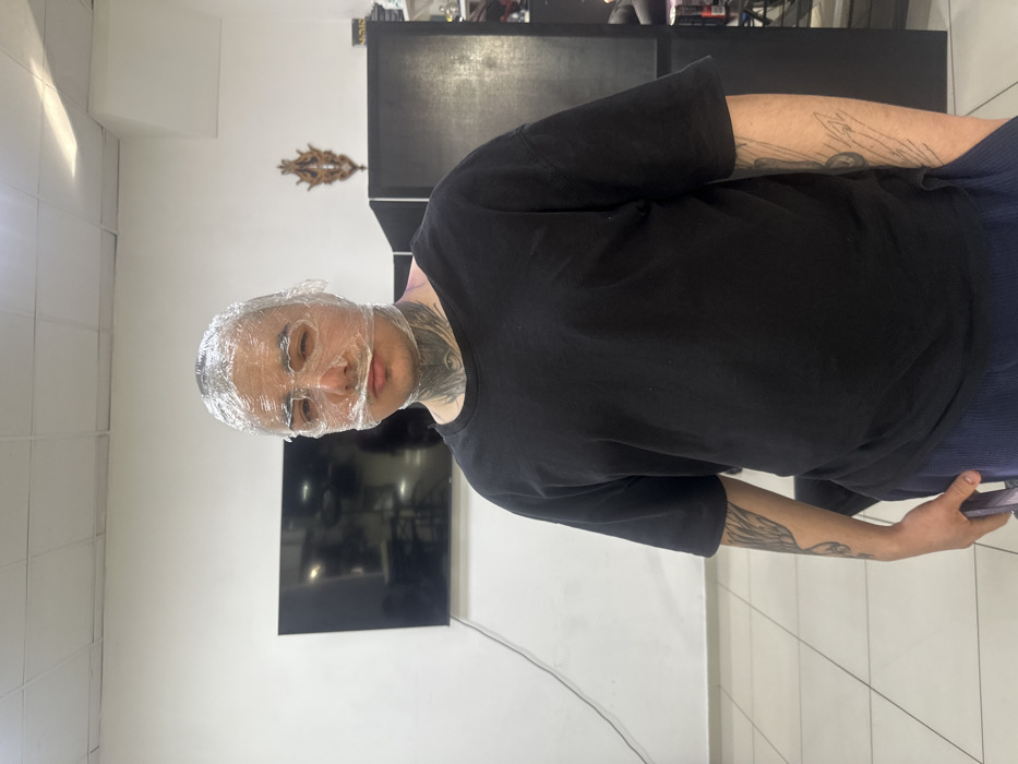 The scene depicts the interior of a studio environment, located in Rostov-on-Don, Russia. A male individual, appearing to be in his 20s or 30s, stands centrally, facing the camera directly. His head and face are entirely wrapped in clear plastic cling film, with cutouts present over the eyes. He has visible tattoos on his neck, including an eye motif, and on both forearms. The individual is wearing a loose-fitting black short-sleeved t-shirt and dark blue ribbed trousers. His left hand is partially obscured, possibly resting in a pocket or holding a small, unseen object. The background consists of plain white walls and a light-tiled floor. To the left, a large, dark-screened flat-panel television is mounted on the wall, with a black cable extending downwards. To the right, a dark, tall storage unit or partition is visible, and above it, an ornate, decorative wooden wall hanging is mounted. The lighting is artificial and evenly distributed throughout the space.