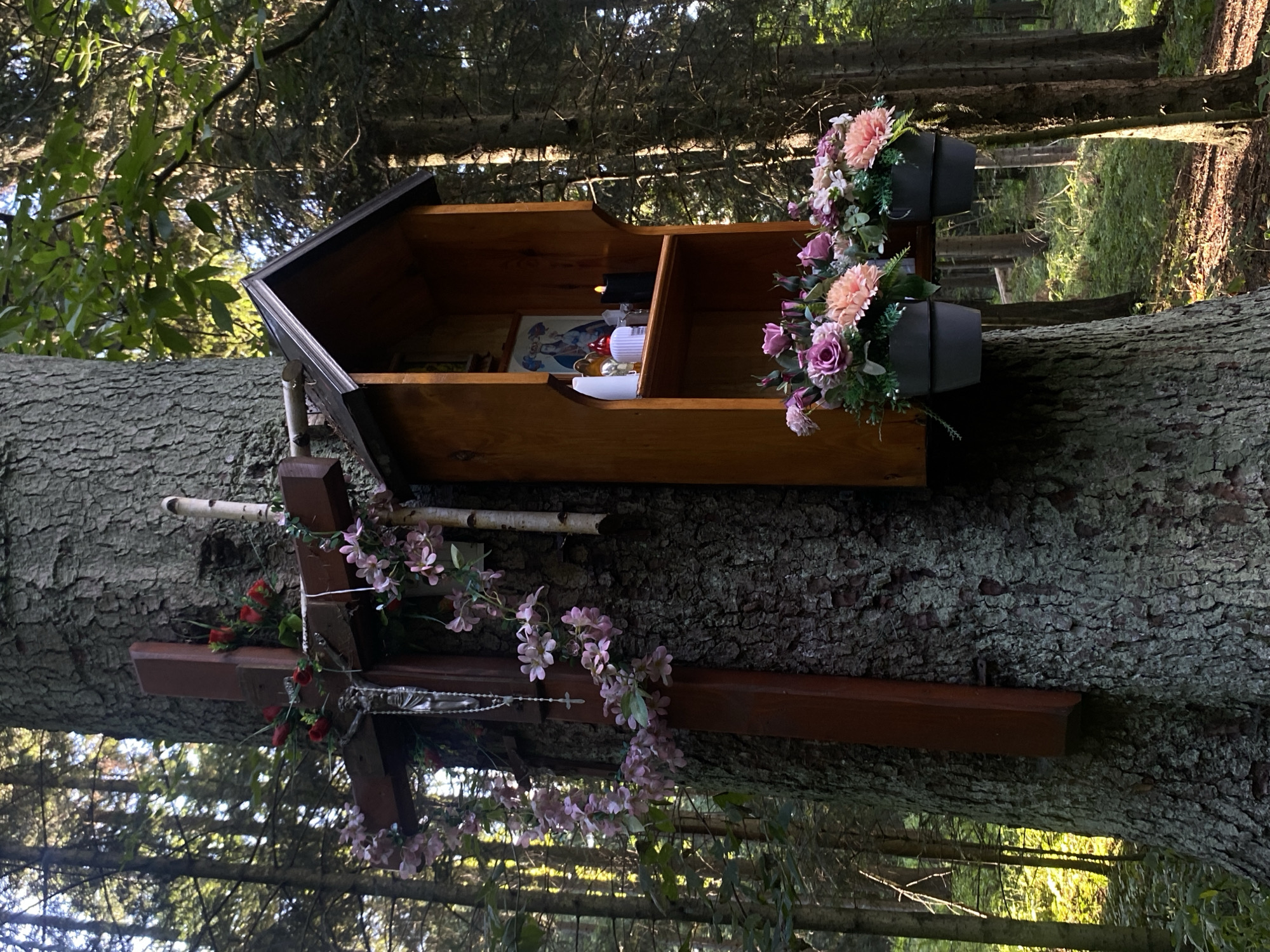 A wooden cross adorned with artificial flowers and a rosary is affixed to a large tree trunk in a forest. To the right of the cross, a wooden shrine is also attached to the tree, containing religious icons, candles, and artificial flowers in pots hanging below. The forest floor is covered with green foliage and dappled sunlight filters through the trees. This scene is located in the city of Stary Sącz, Poland, near the Franciscan Monastery.
