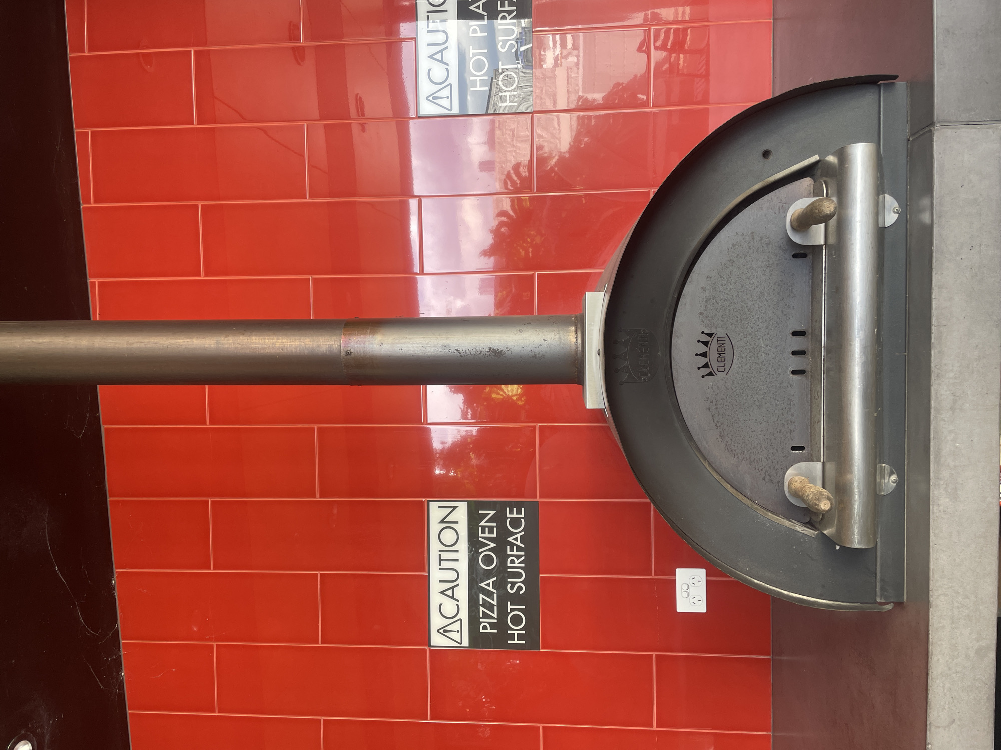 The image shows a close-up of a pizza oven in a commercial setting. The oven is dark grey and arched, with a metal flue pipe extending vertically upwards. Two wooden handles protrude from the front of the oven. On the red tiled wall behind the oven, there are two cautionary signs. The sign on the left reads "CAUTION PIZZA OVEN HOT SURFACE." The sign on the right is partially obscured but appears to read "CAUTION HOT PLAT HOT SURF." Below the left sign, a white electrical outlet with three sockets is visible on the red tiled wall. The overall environment appears to be an outdoor or semi-outdoor area, possibly a restaurant patio or food stall, given the red tiled backsplash and the type of equipment. The image does not provide enough context to determine its specific location within the city of Brisbane, Australia. There are no people visible in the frame.