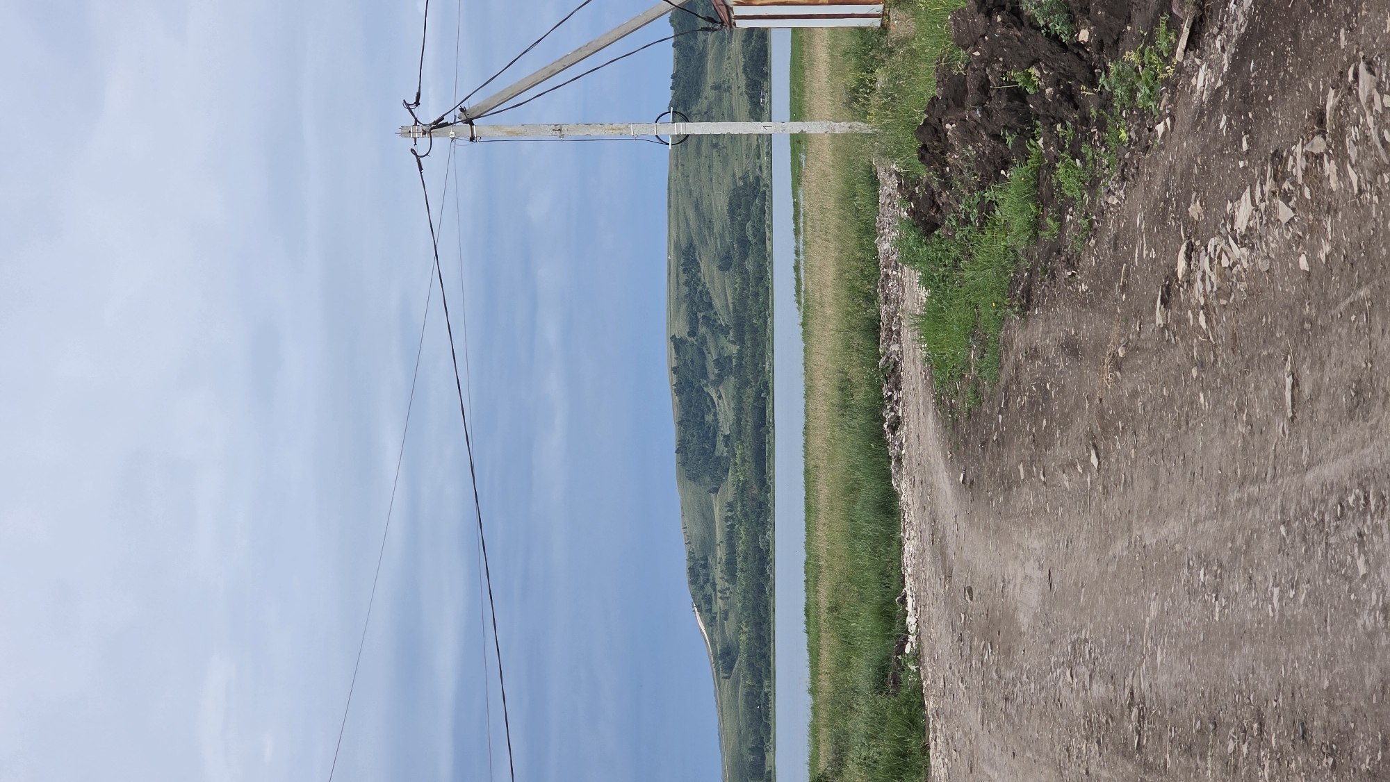 The image depicts a rural landscape near Kupoyarovo, Russia, dominated by a dirt road in the foreground leading towards a body of water. A power pole with visible wires stands on the right side of the road, adjacent to a small, partially visible structure with white and reddish-brown siding. Beyond the road and a strip of tall grass, a wide, calm body of water stretches across the midground. In the background, a rolling, verdant hill covered in trees and patches of grass rises against a cloudy, overcast sky. The overall scene suggests a quiet, natural setting.