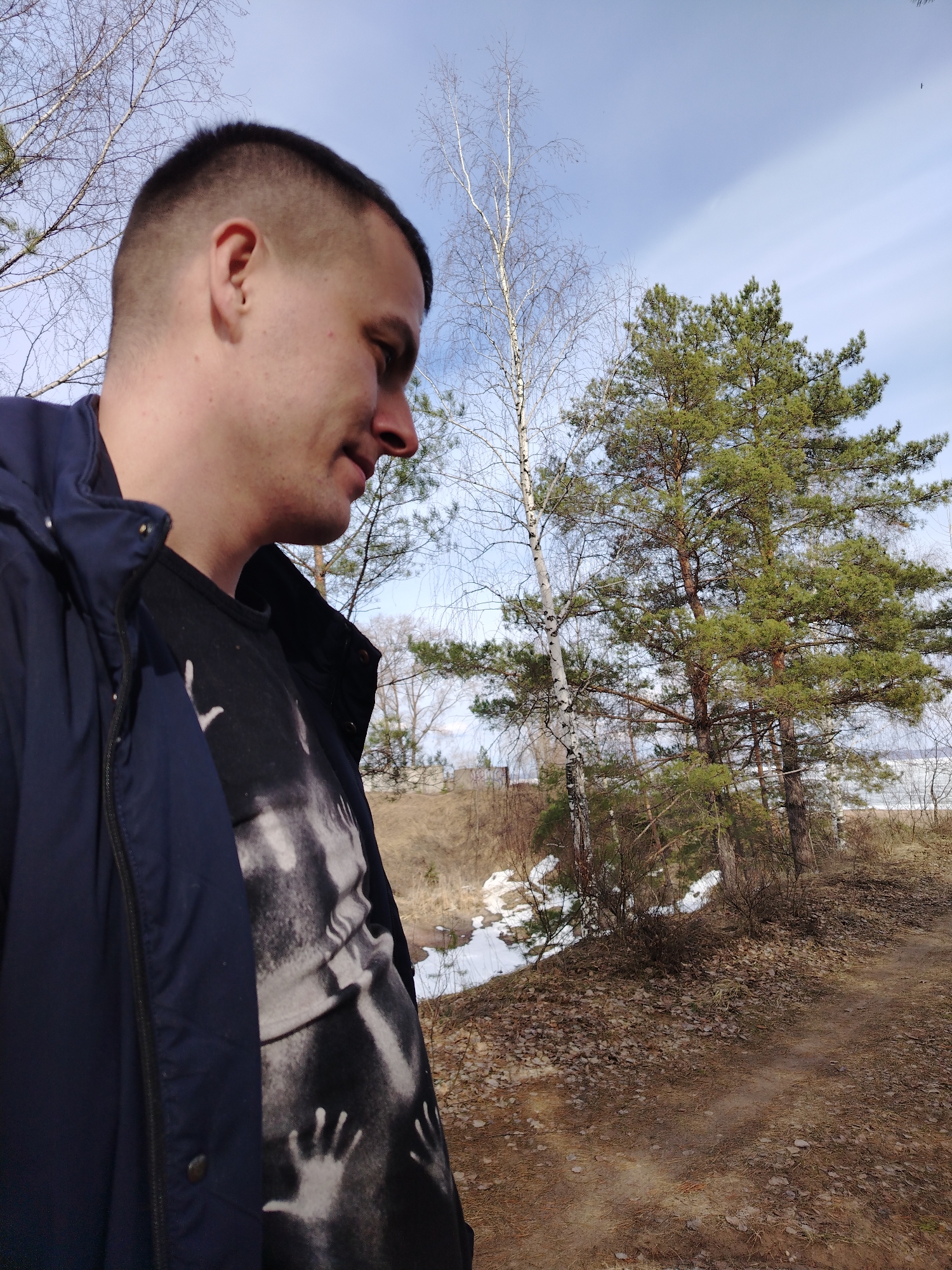A man with a short haircut stands in an outdoor location, likely a wooded area or park. He is wearing a dark blue jacket and a black t-shirt with a graphic design. He is looking down and to his right.
Behind him, the landscape includes trees, mostly deciduous with some coniferous, and patches of snow on a hillside. The ground is covered in leaves and bare earth. In the background, there is a glimpse of water or a body of water and sky with scattered clouds.
The location is in Noviy, Russia, based on possible identification of the setting as a landscape characteristic of the region. The light suggests it is daytime, possibly in the late morning or early afternoon.