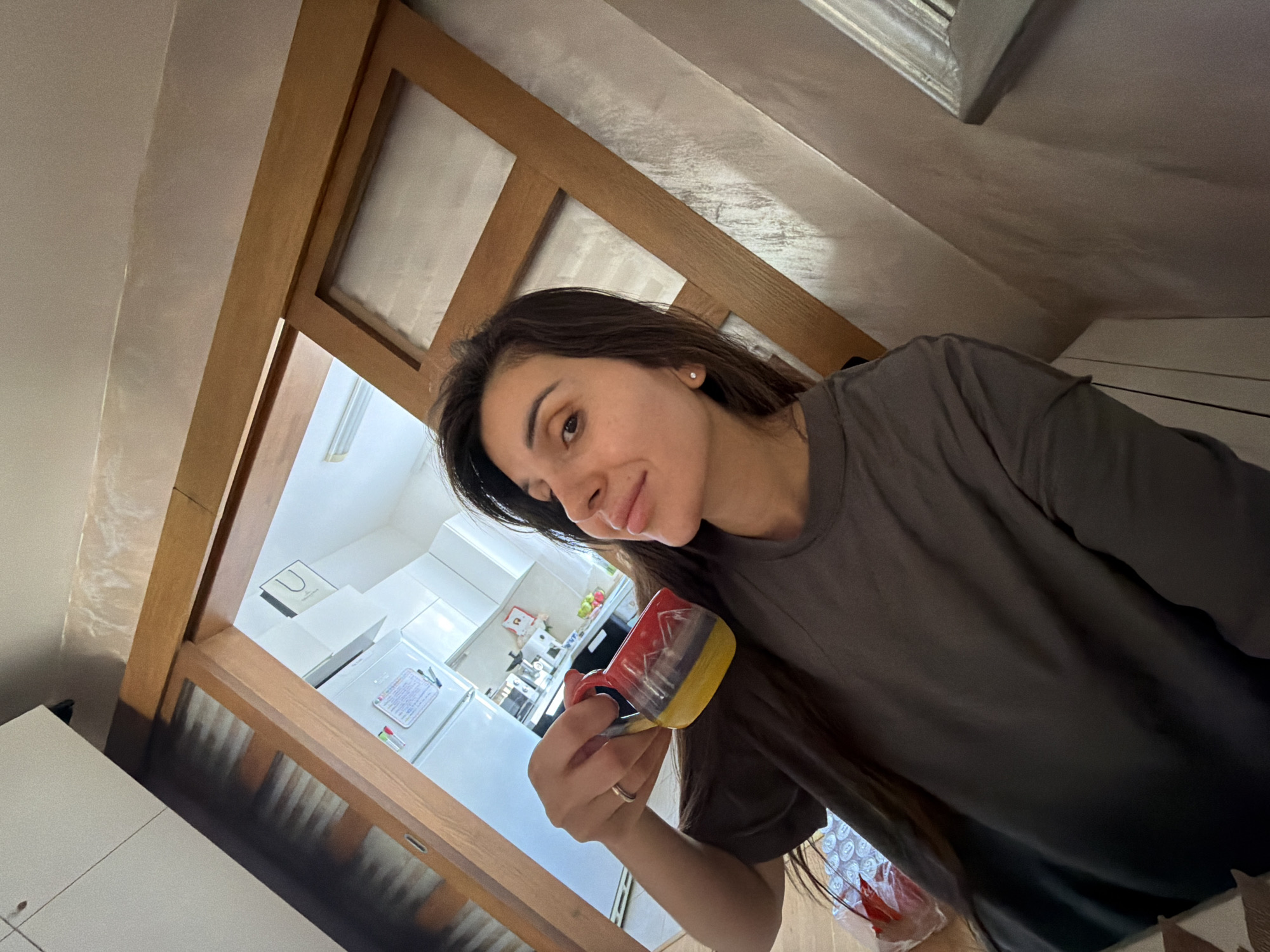 A woman is indoors, holding a colorful mug, possibly in an apartment or residence in Budva, Montenegro. The mug has red, purple, and yellow colors. A kitchen with white cabinets and appliances, including a refrigerator, is visible in the background through a wooden-framed doorway. The woman is wearing a dark gray t-shirt. The setting appears to be a casual domestic scene.