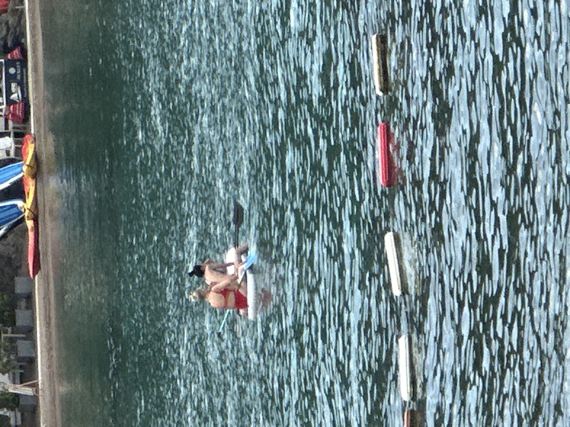 The image depicts a serene, sun-drenched day at a beach, likely in Budva, Montenegro. The water, a calm expanse of deep blue-green, reflects the bright sky above, creating a shimmering surface. In the mid-ground, two individuals, likely women, are enjoying a paddleboarding excursion. One is seated on a white paddleboard, wearing a red bikini and with blonde hair tied up. The other sits behind her, her dark hair pulled back, and appears to be wearing a dark top. A paddle, predominantly blue, is visible in the water between them. The background reveals the sandy shore of the beach, lined with what appear to be potted plants and some beach equipment, including colorful kayaks or rafts. Further in the distance, some structures are visible, hinting at the vibrant resort town of Budva. In the foreground, several white and red floating buoys are sparsely scattered across the water, marking a boundary or area within the bay. The overall atmosphere is one of leisure and relaxation.