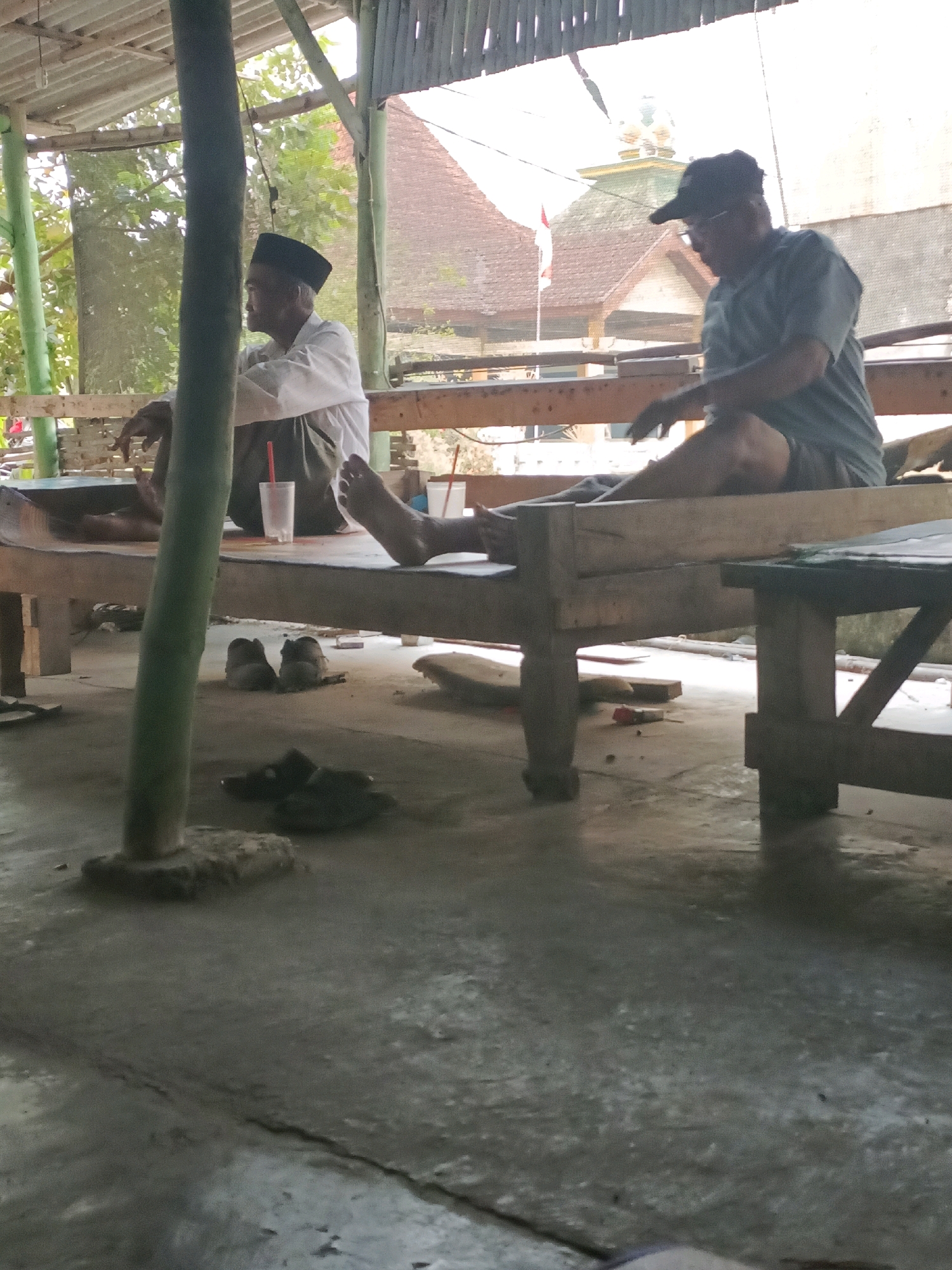 Two men are seated on a wooden bench under a partially open-roofed structure. The man on the left, wearing a white shirt and a black skullcap, is facing to the left. The man on the right, wearing a grey t-shirt, shorts, and a baseball cap, is looking down and appears to be holding something in his right hand. A white cup with a red straw is visible on the bench between them. In the foreground, on the concrete floor, are several pairs of sandals and a large cylindrical concrete base supporting a wooden pole. The background shows a building with a red tiled roof and trees. The scene appears to be outdoors in a relaxed setting, possibly in Rancang Kencono, Indonesia.