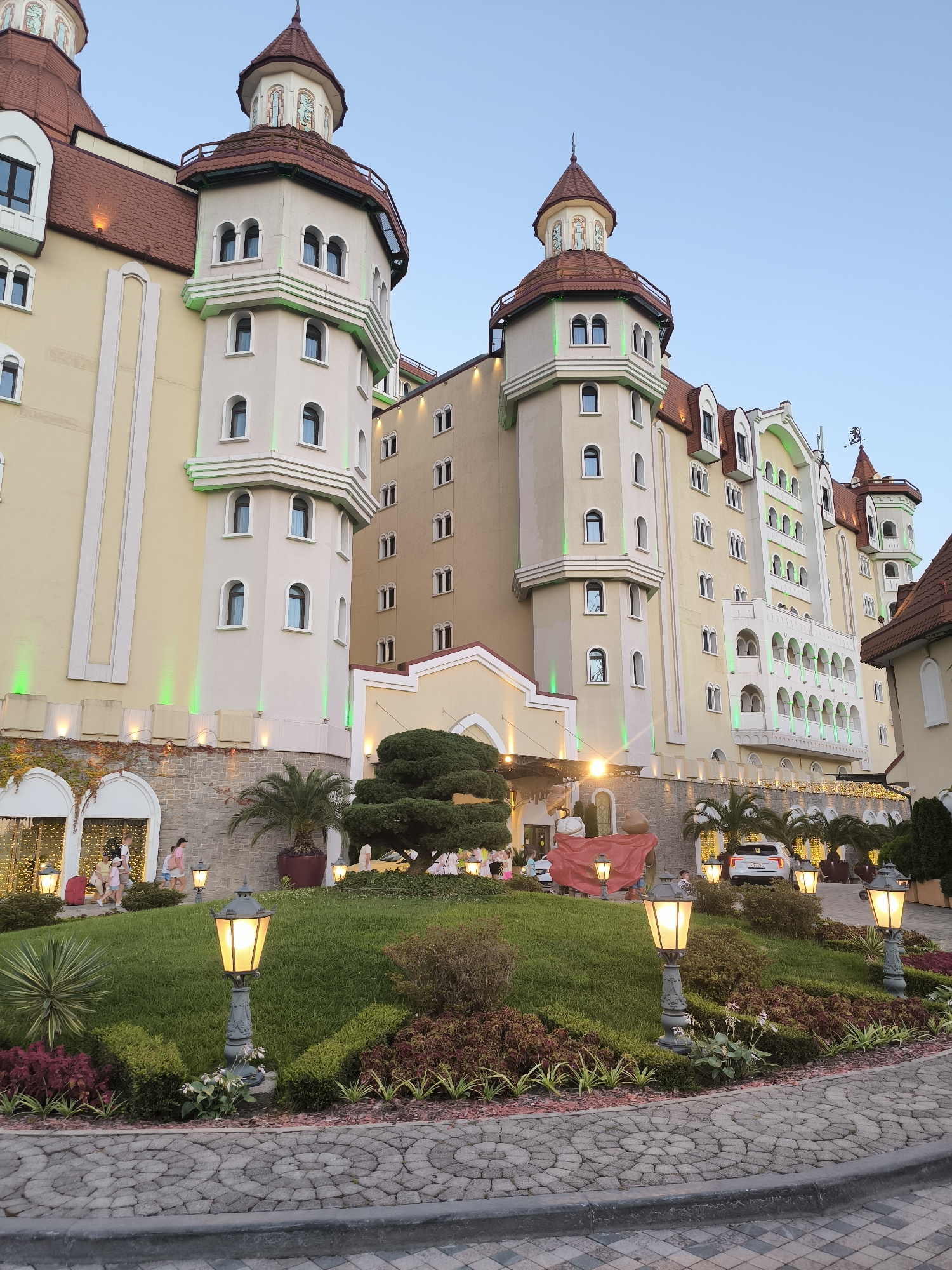 The image shows a large, multi-story building designed in a fairy-tale castle style, located in the city of Sirius, Russia. The building features multiple towers with red conical roofs, arched windows, and decorative crenellations. It is painted in light yellow and beige tones with accents of green lighting illuminating the lower levels of the towers. In the foreground, a landscaped garden area is visible, with a cobblestone pathway leading through it. Several ornate lampposts with lit bulbs are placed along the pathway and in the garden. People are seen walking in the background near the entrance of the building. A white car is parked in front of the building. The sky is clear and blue, indicating it is likely daytime.