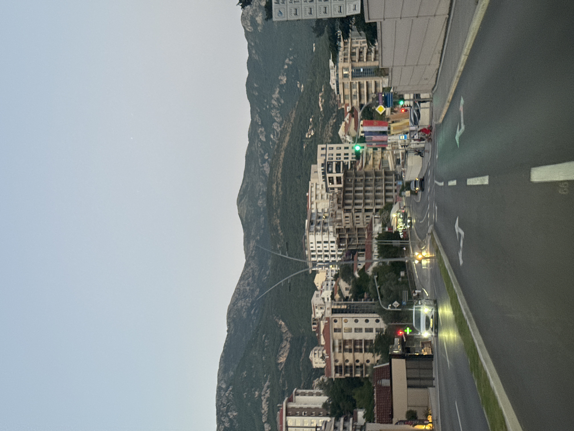 The image captures a street scene in Boreti, Montenegro, during dusk. The foreground shows a paved road with white lane markings, including an arrow indicating a left turn. A white car is visible in the distance, approaching an intersection with traffic lights. The background features a densely populated urban area with numerous multi-story buildings of varying architectural styles, some appearing modern with glass facades and others with traditional red-tiled roofs. Beyond the buildings, a lush green mountain range dominates the horizon, its slopes partially obscured by the twilight haze. The sky is a pale, clear blue, transitioning to a darker hue towards the top of the frame. The overall impression is of a coastal town nestled between the mountains and the sea, with the subtle glow of streetlights and building lights beginning to appear as nightfall approaches.