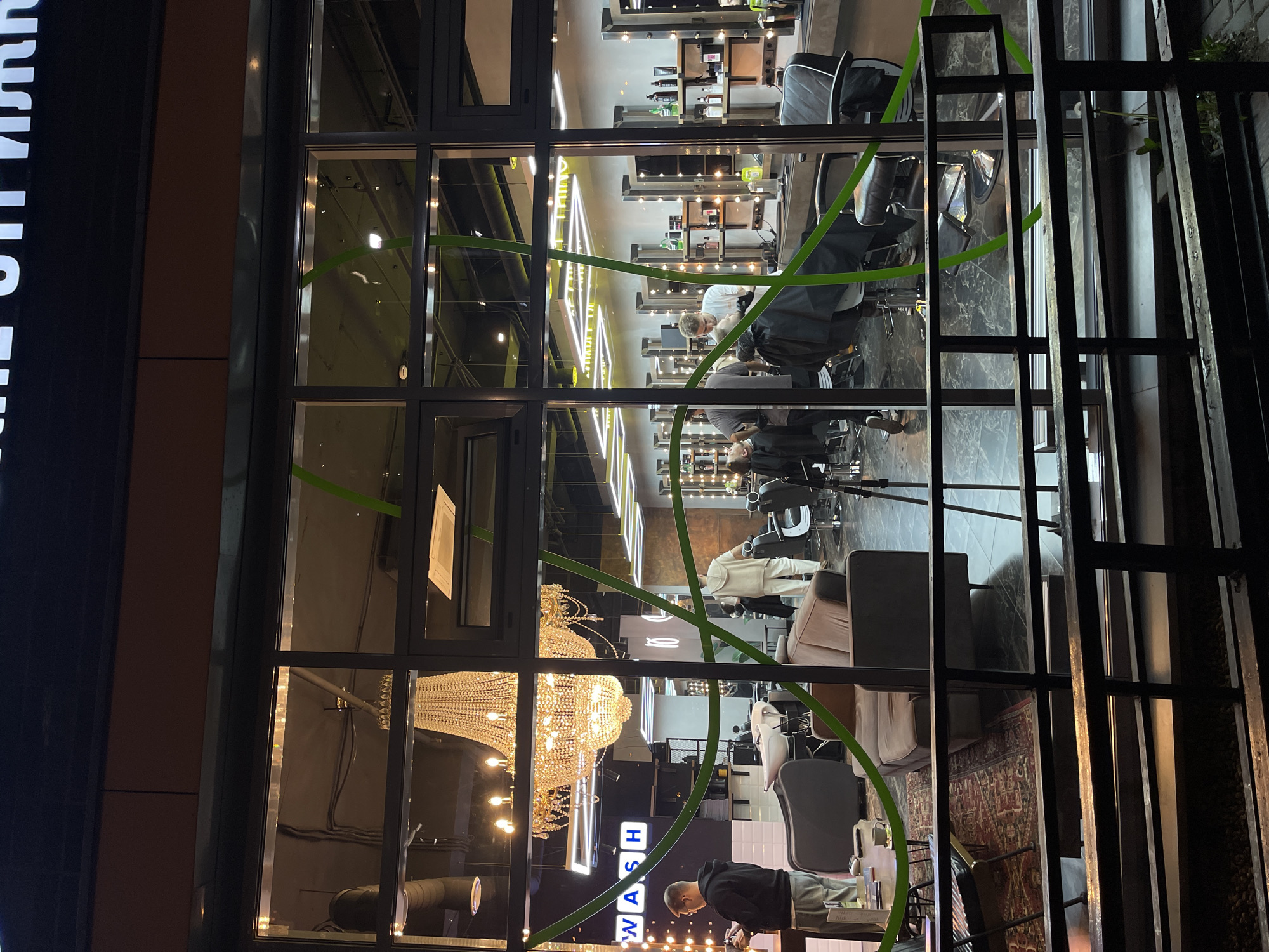 A nighttime view through a large glass window reveals the interior of a barbershop in Khimki, Russia. The interior is brightly lit with a chandelier and neon lights. Several people are visible: barbers are working on clients seated in barber chairs. One person is seen at the front of the shop appearing to clean. The shop is equipped with styling stations, mirrors, and waiting area seating. Green decorative vines are visible on the exterior of the window. A railing is seen to the right of the window.