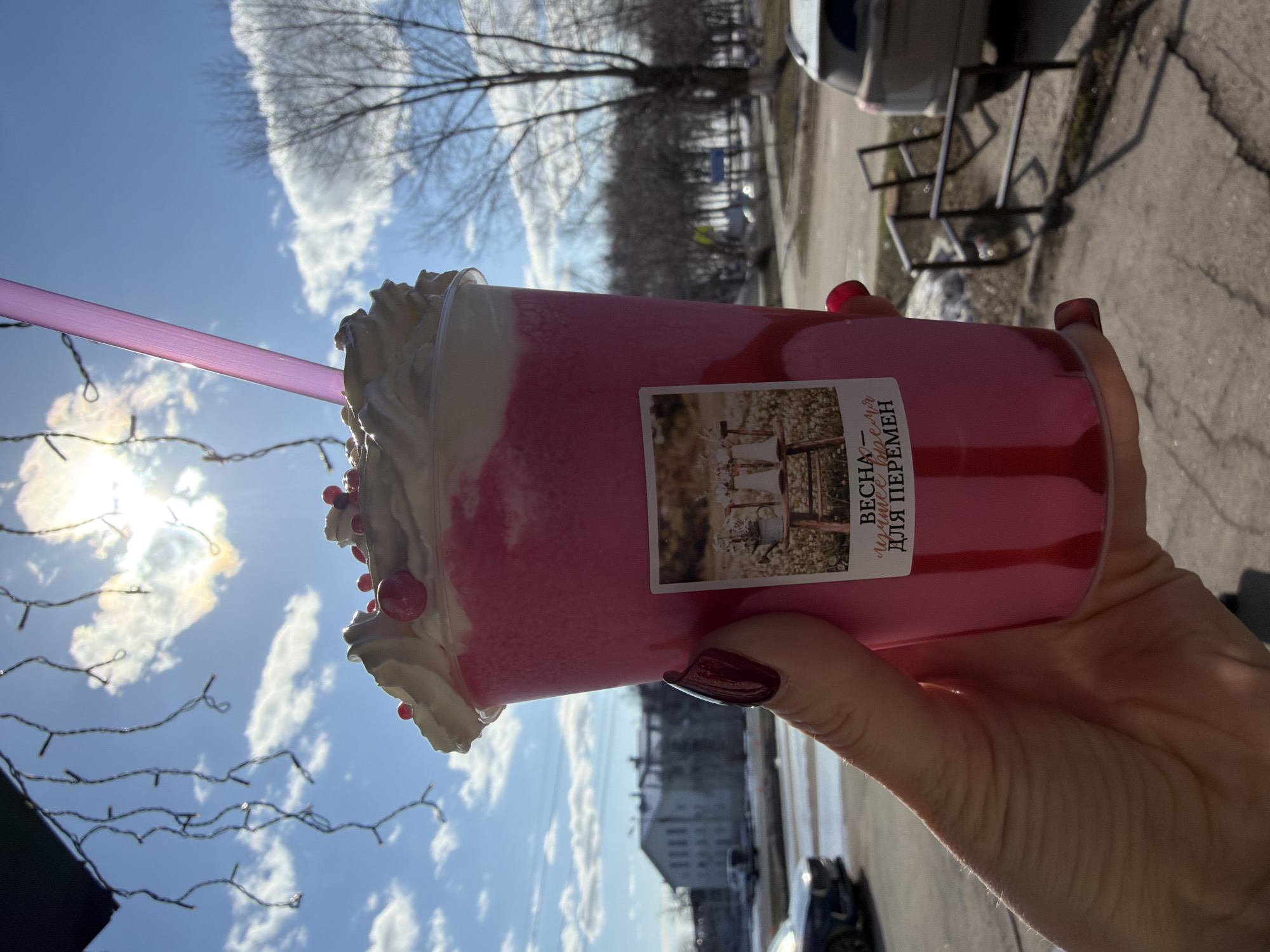 A photograph shows a person's hand holding a pink beverage with whipped cream and a pink straw outdoors. The beverage has a label with the words "ВЕСНА - лучшее для ПЕРЕМЕН" written in Russian. The person's nails are painted dark red. The background features a city scene with buildings, trees, and a partly cloudy sky. The presence of what appear to be bare trees suggests a spring or autumn season. Based on the text on the label, the location is likely Votkinsk, Russia.
