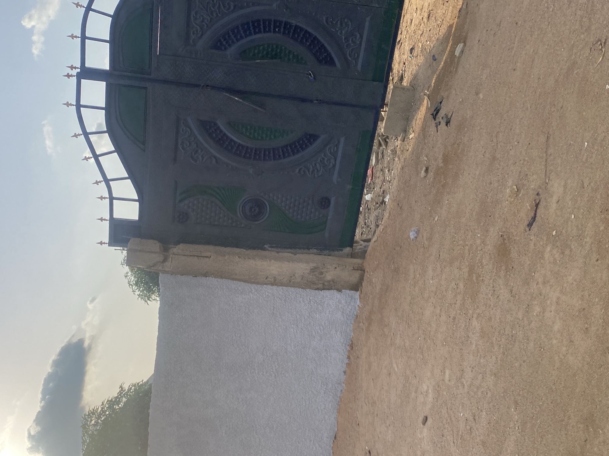 The image displays a closed, dark metal gate with intricate circular patterns and green accents, mounted on a light-colored wall. The gate has a curved top with decorative spikes. To the right of the gate, there is a dirt path littered with debris. A rough, light-colored wall extends from the left side of the gate. Above the gate and wall, the sky is visible, with some clouds and a bright light suggesting it is daytime, likely late afternoon due to the warm tones. The scene appears to be outdoors in an unpaved area. There is no text visible in the image.