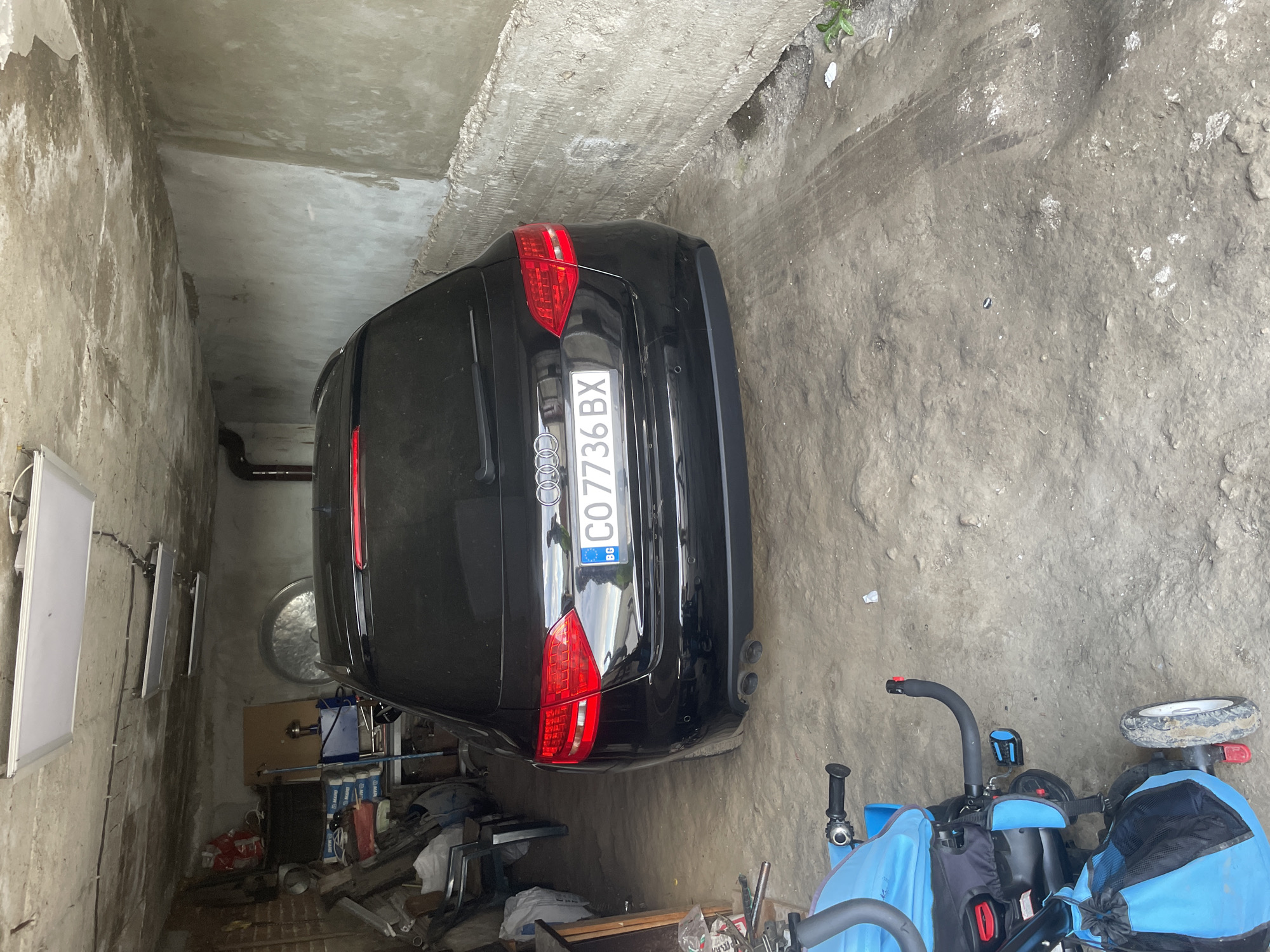 The image displays the rear of a black Audi car parked within a concrete garage or storage area. The car, an SUV or wagon model, is positioned on a rough, uneven concrete and dirt floor, which shows tire marks. Its license plate, "C07736 BX," is clearly visible, along with the "BG" country code, indicating Bulgaria. The car's red taillights, parking sensors on the bumper, and dual exhaust pipes on the right are prominent.
To the lower right of the car, a blue and black child's tricycle or stroller is parked, with one of its wheels appearing muddy. The surrounding walls and ceiling are composed of unfinished, raw concrete, exhibiting a weathered appearance. Along the left concrete wall, several whiteboards or panels are mounted, accompanied by visible electrical wiring. In the background, behind and beneath the car, a cluttered storage area contains various items, including boxes, pipes, and bottles or containers, some labeled with the brand "Mapei." A circular vent or window is also discernible on the back wall. The scene is illuminated by ambient daylight, suggesting an indoor setting during the daytime. The photograph captures a static moment of stored items in Ihtiman, Bulgaria.